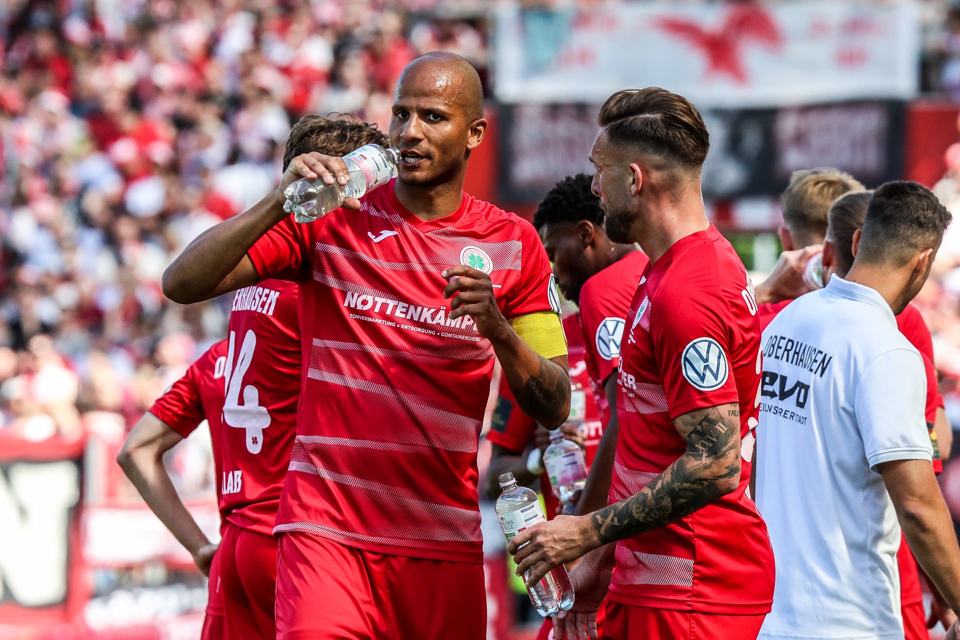 [{"clang_id":1,"value":"Fußballspieler in roten Trikots feiern auf dem Spielfeld. Im Hintergrund eine volle Zuschauertribüne. Ein Spieler hält eine Wasserflasche hoch."}]