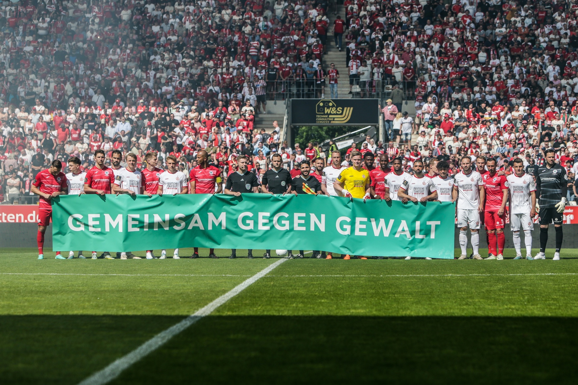 [{"clang_id":1,"value":"Fußballstadion: Spielerteams mit Banner \"Gemeinsam gegen Gewalt\". Im Hintergrund dicht gedrängte Zuschauermenge in rot-weißer Kleidung."}]
