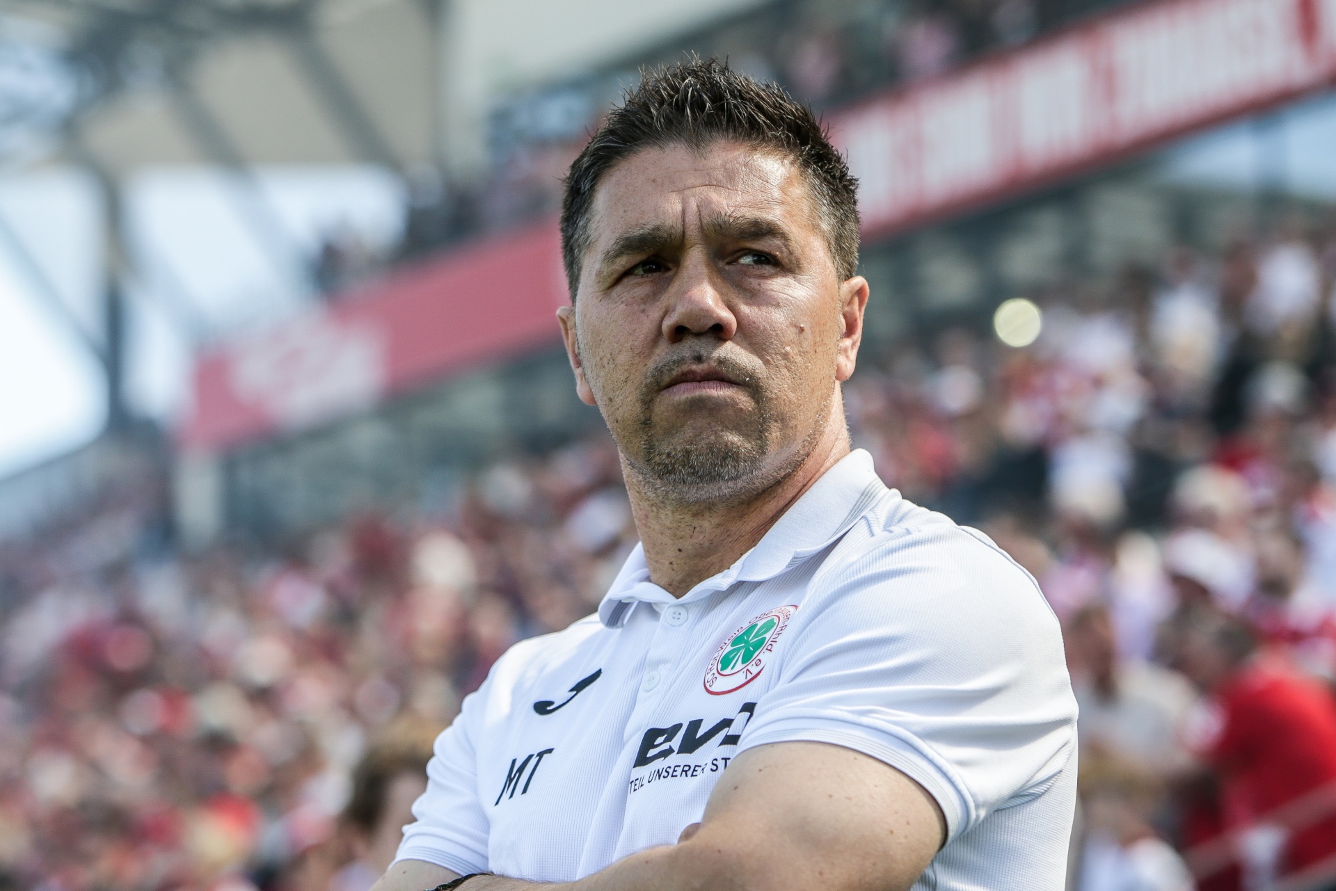 [{"clang_id":1,"value":"Mann mit kurzgeschorenen Haaren und Bart, trägt weißes Trikot mit grünem Logo und Werbeaufdruck. Hintergrund: verschwommene Menschenmenge und Stadion."}]