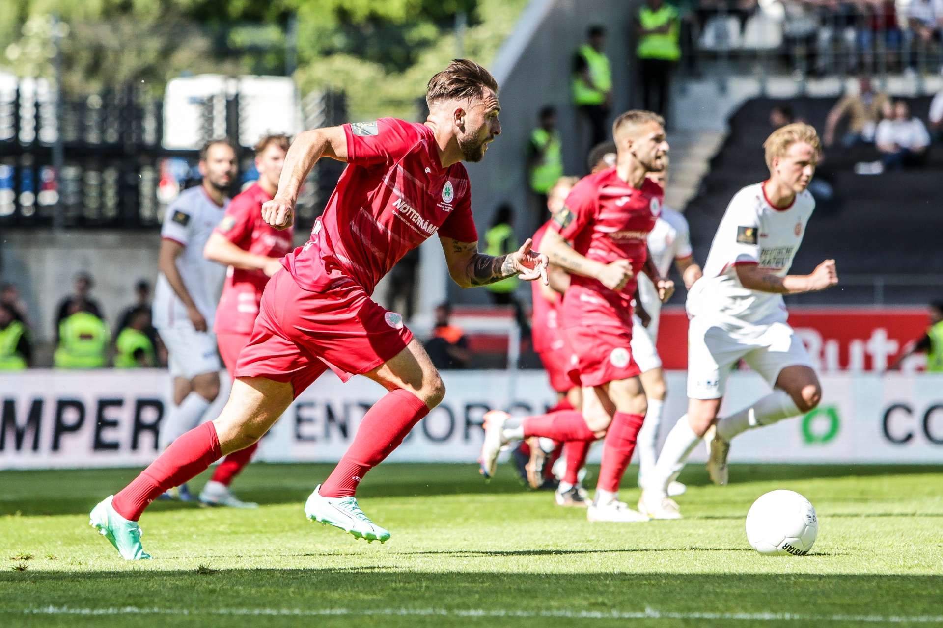 [{"clang_id":1,"value":"Fußballspiel: Spieler in roten Trikots laufen auf grünem Rasen auf den Ball zu. Im Hintergrund Zuschauer und Stadiontribünen."}]