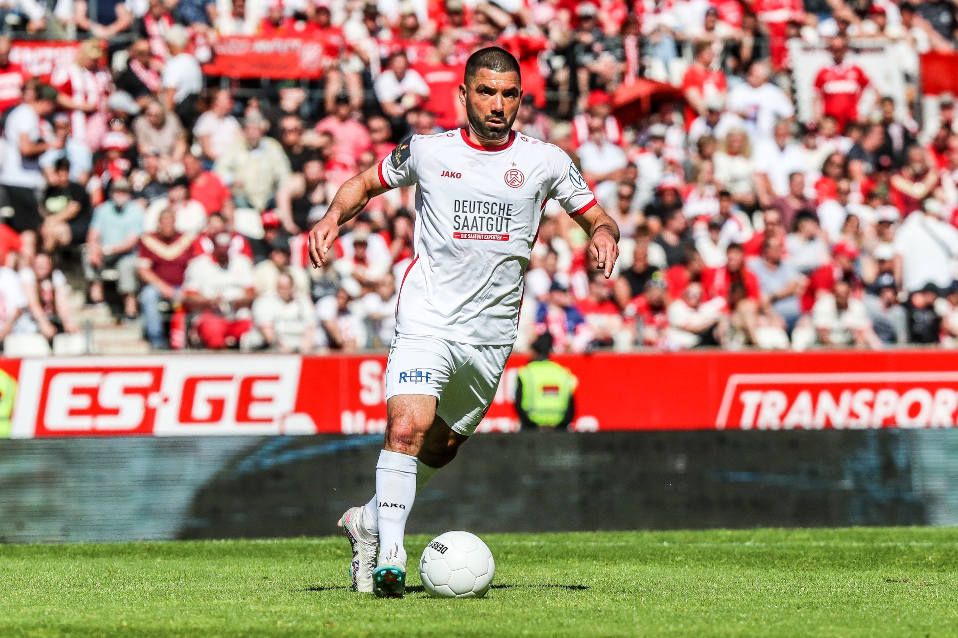 [{"clang_id":1,"value":"Fußballspieler in weißem Trikot mit Vereinslogo auf grünem Rasen, Hintergrund voller Zuschauer in rot-weiß. Reklamebanner im Hintergrund."}]
