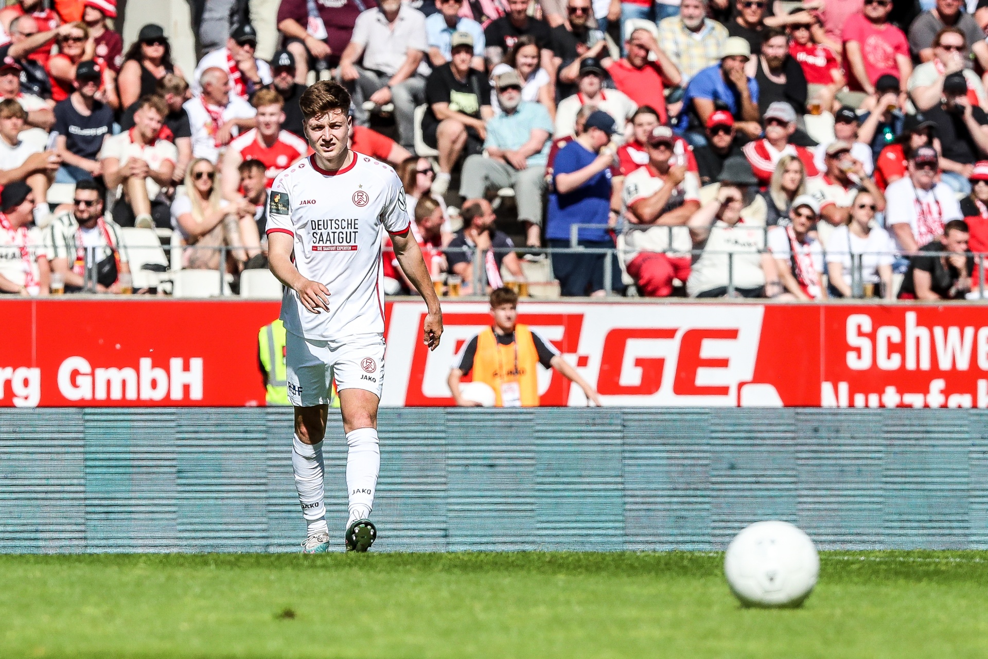 [{"clang_id":1,"value":"Fußballspieler mit weiß-rotem Trikot steht auf grünem Rasen vor Zuschauermenge. Im Hintergrund Werbebanner. Ein Fußball liegt am Boden."}]