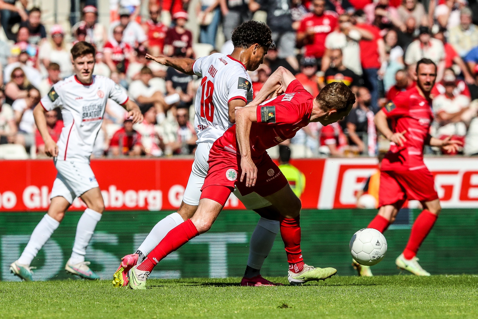 [{"clang_id":1,"value":"Fußballspiel: Zwei Spieler in rot und weiß kämpfen um den Ball auf grünem Rasen. Im Hintergrund Zuschauerreihen."}]