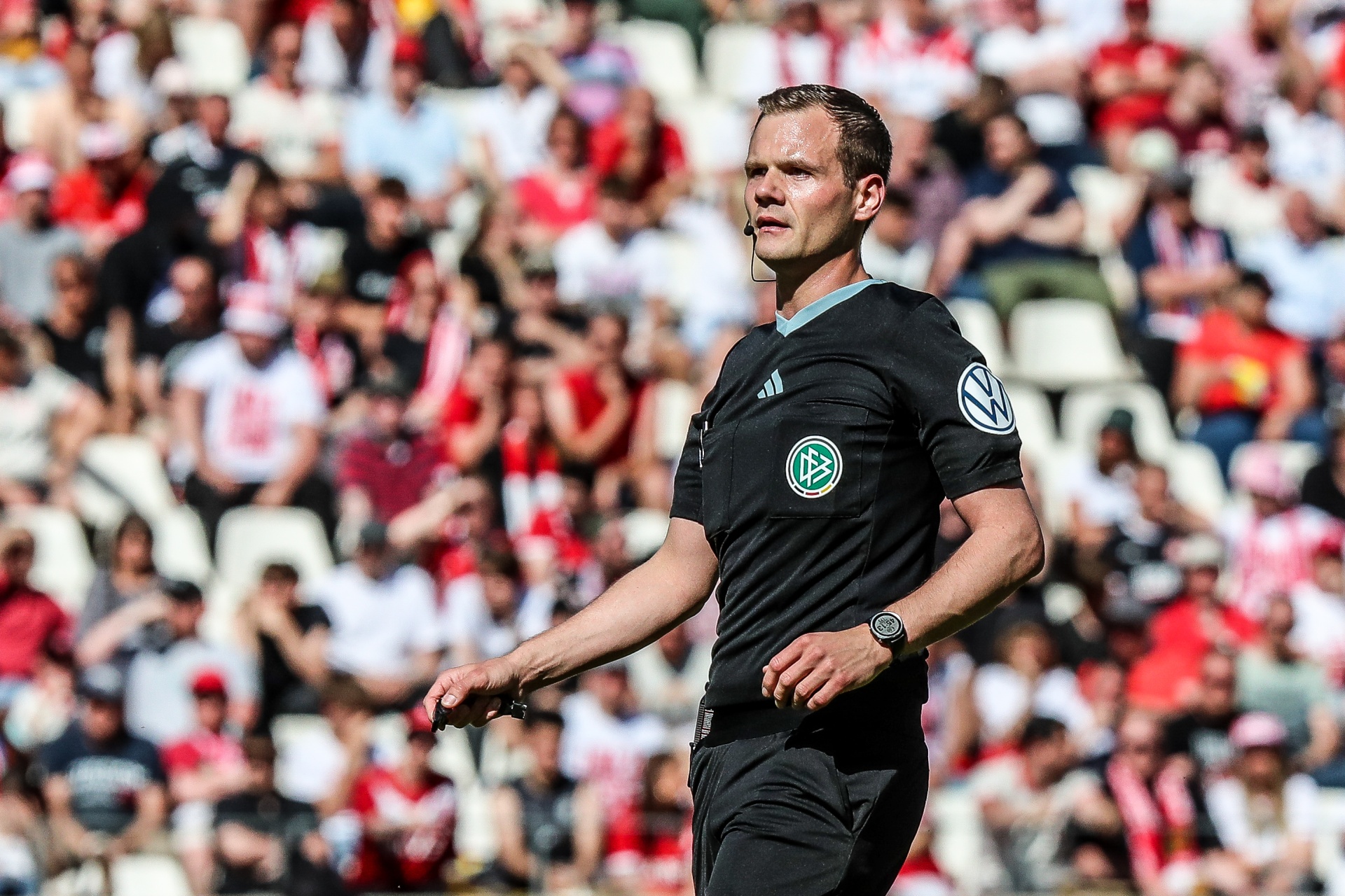 [{"clang_id":1,"value":"Fußballschiedsrichter in schwarzer Uniform mit weißen Ärmeln steht auf dem Spielfeld vor einer Zuschauermenge in rot-weißer Kleidung."}]