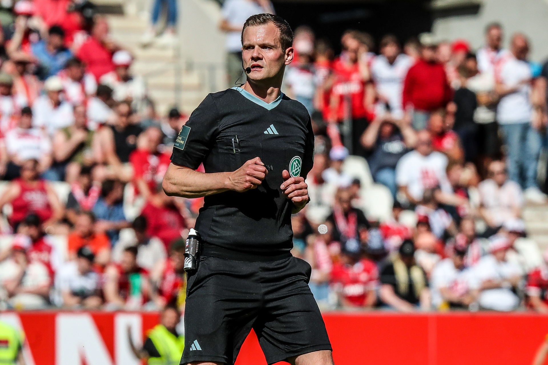 [{"clang_id":1,"value":"Fußballschiedsrichter in schwarzer Sportkleidung mit Adidas-Logo rennt auf dem Spielfeld vor einer Zuschauermenge mit rot-weißer Kleidung."}]