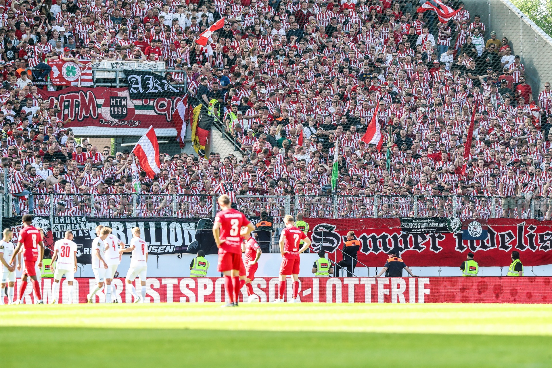 [{"clang_id":1,"value":"Fußballstadion: Spieler stehen vor voll besetzter Tribüne mit rot-weißen Fahnen und Banner. Im Hintergrund Spielrasen."}]