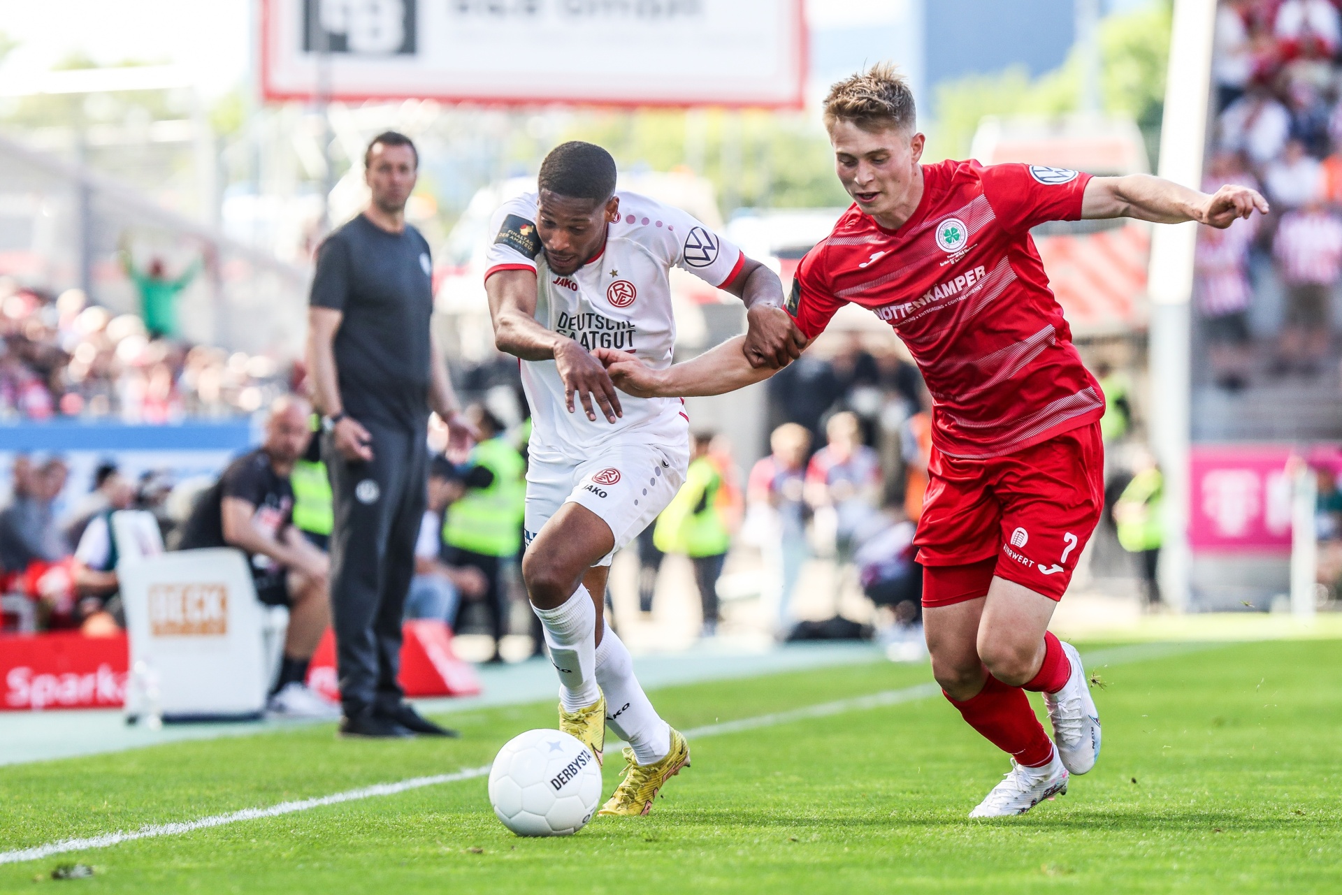 [{"clang_id":1,"value":"Zwei Fußballspieler im Wettkampf. Links ein Spieler mit rotem Trikot und braunen Haaren, rechts ein Spieler mit rotem Trikot und blonden Haaren. Beide kämpfen um den Ball auf grünem Rasen. Zuschauer im Hintergrund."}]