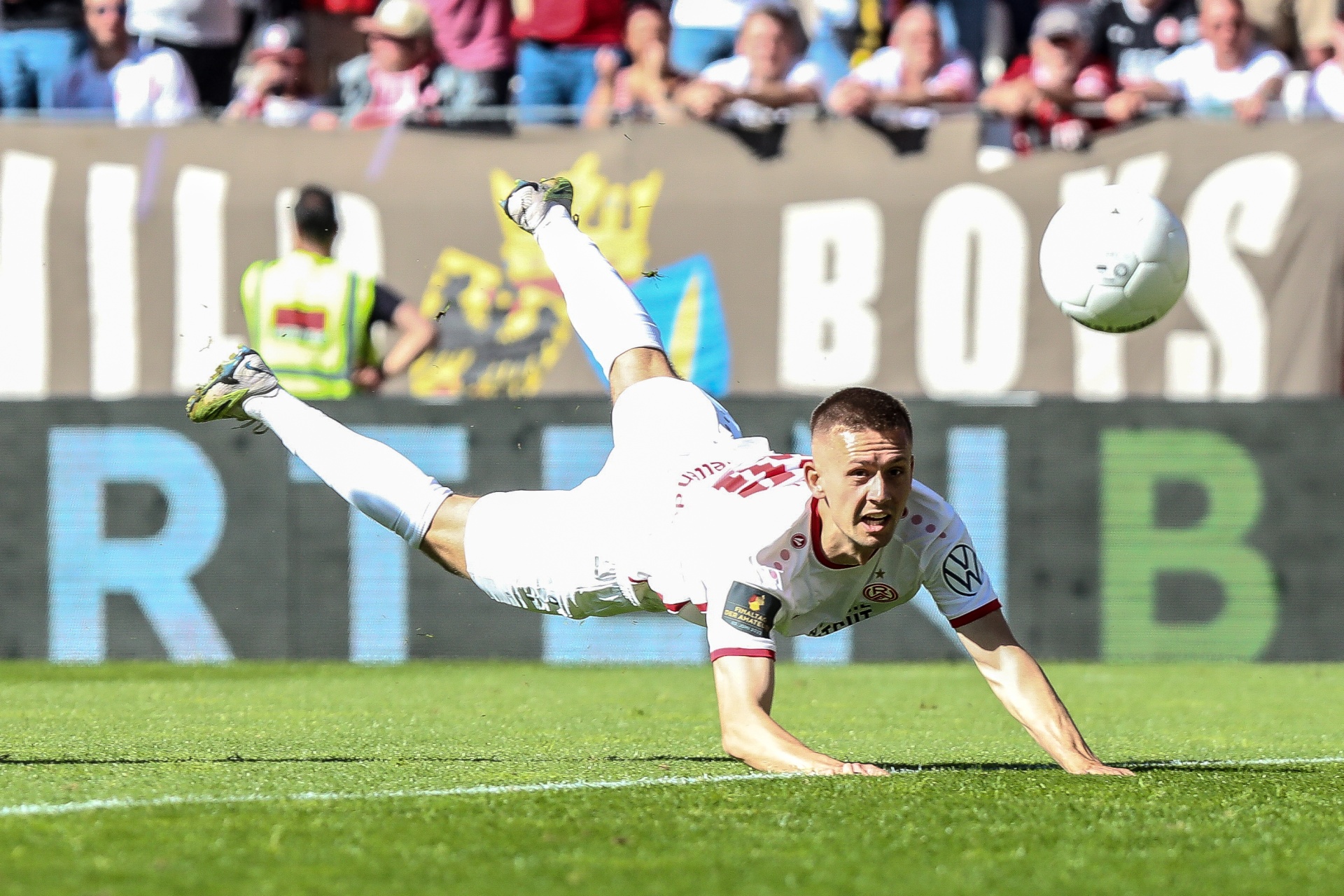 [{"clang_id":1,"value":"Fußballspieler in Rot-Weiß rutscht auf grünem Rasen, Arme vorgelegt, ein Bein hoch erhoben, der Ball in der Luft. Im Hintergrund Zuschauer und Banner."}]
