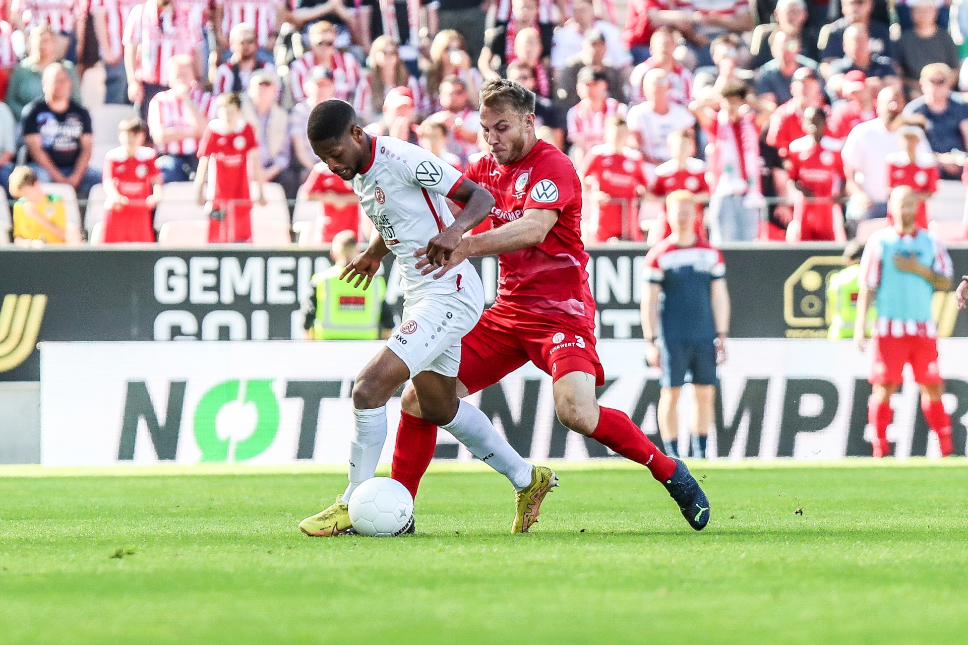 [{"clang_id":1,"value":"Zwei Fußballspieler im Spiel. Ein Spieler mit weißem Trikot und der andere mit rotem Trikot kämpfen um den Ball auf dem grünen Rasen. Im Hintergrund Zuschauer."}]