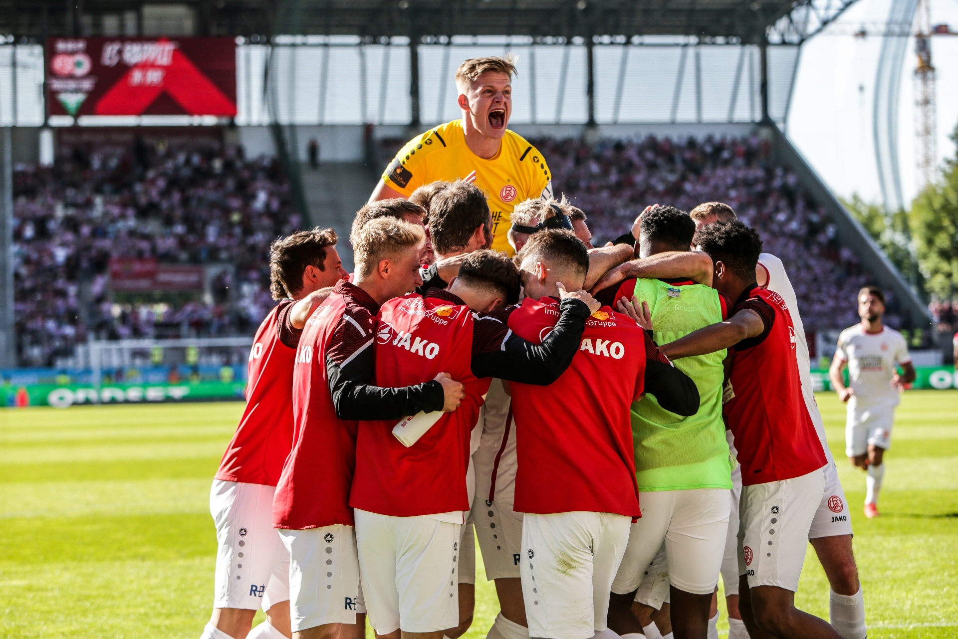 [{"clang_id":1,"value":"Fußballspieler in roten Trikots jubeln eng umschlungen auf dem Spielfeld. Im Hintergrund Zuschauer auf den Tribünen."}]