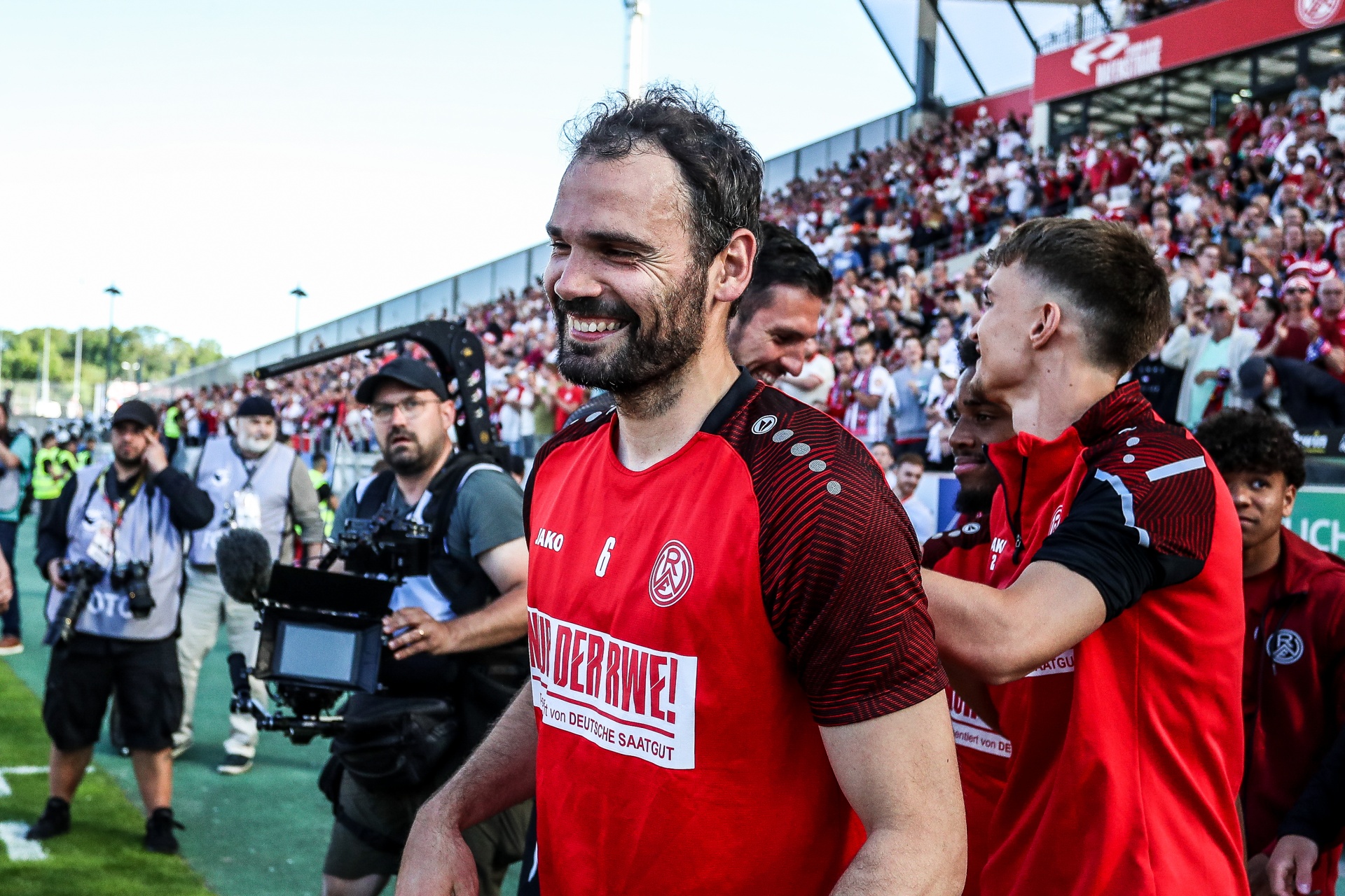 [{"clang_id":1,"value":"Zwei Fußballspieler in roten Trikots, einer lächelt in die Kamera, werden von jubelnden Zuschauern umgeben. Im Hintergrund ein Fußballstadion und eine Kamera."}]