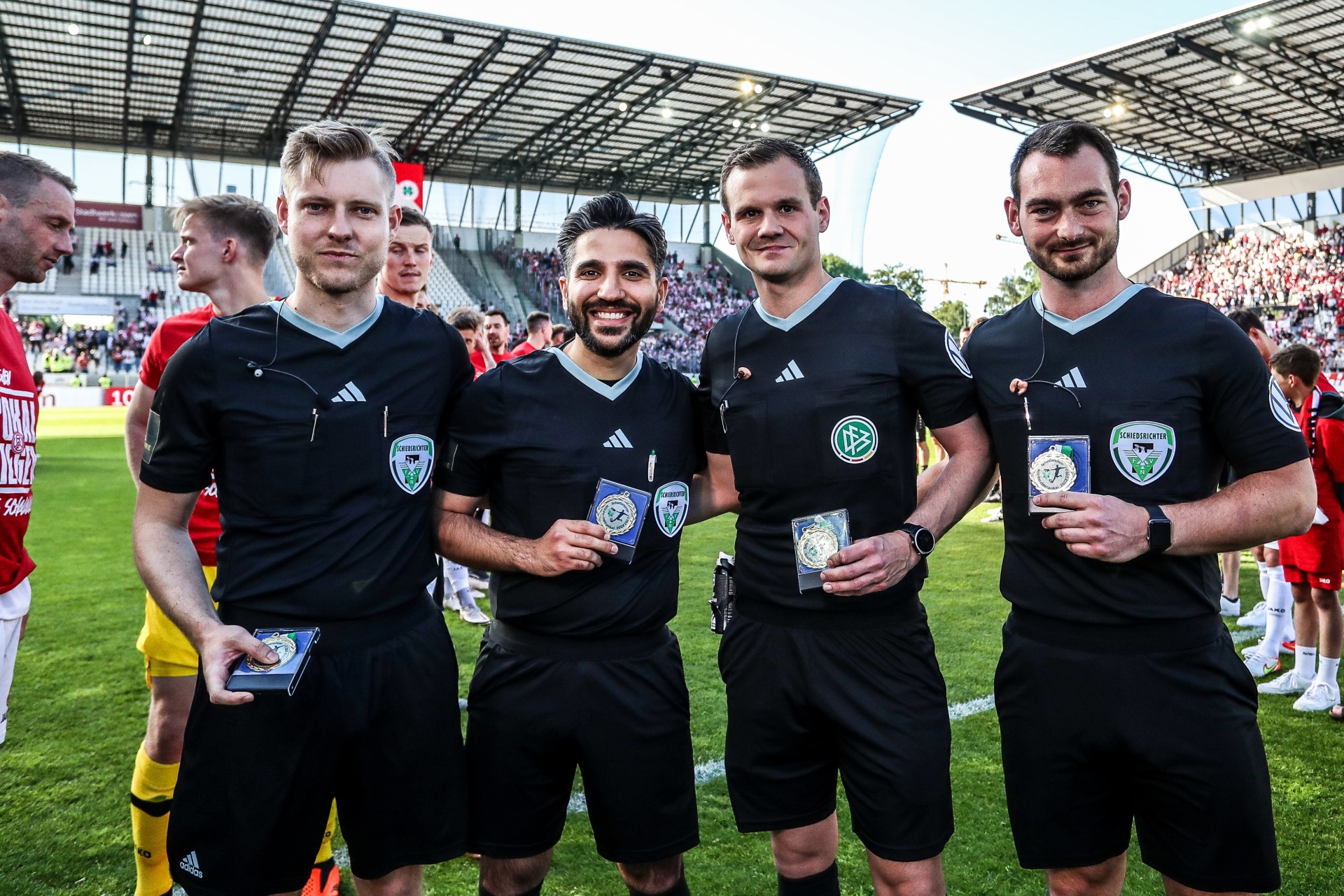 [{"clang_id":1,"value":"Vier Schiedsrichter in schwarzer Sportkleidung mit Vereinslogo stehen lächelnd auf einem Fußballfeld vor Zuschauern und Stadion. Je einer hält eine kleine Auszeichnung."}]
