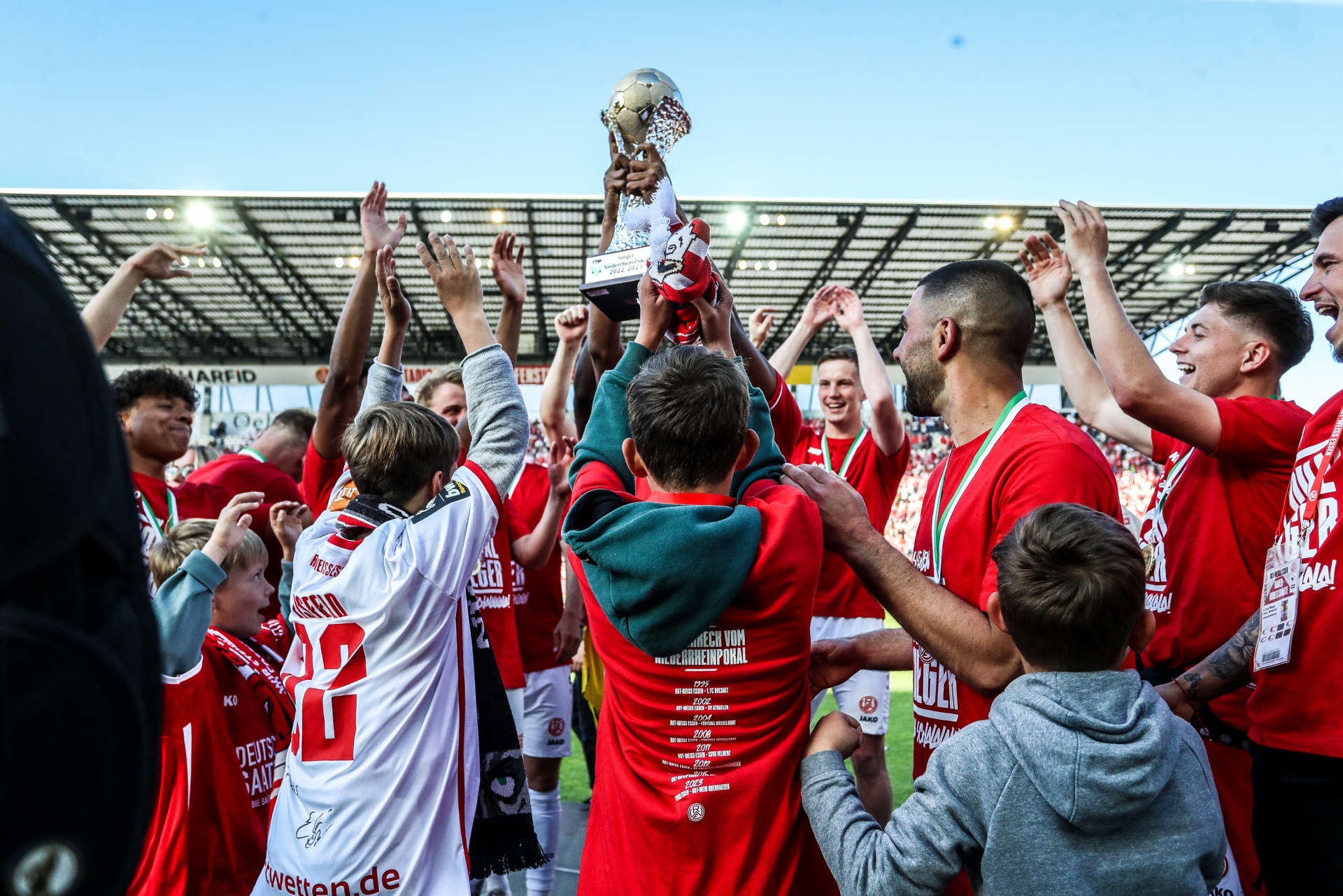 [{"clang_id":1,"value":"Fußballmannschaft feiert Sieg. Spieler in rot-weißen Trikots heben eine goldene Trophäe in die Luft, umgeben von jubelnden Fans im Stadion."}]