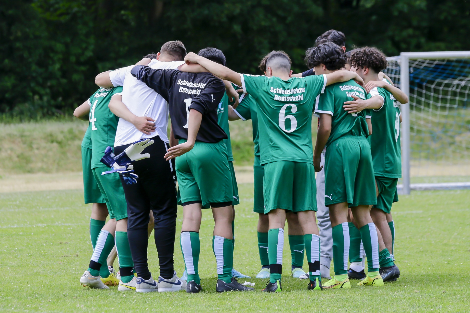 [{"clang_id":1,"value":"Fußballteam im Kreis. Spieler tragen grüne Trikots und schwarze Shorts, ein Trainer steht dabei. Hintergrund: Fußballfeld und Bäume."}]