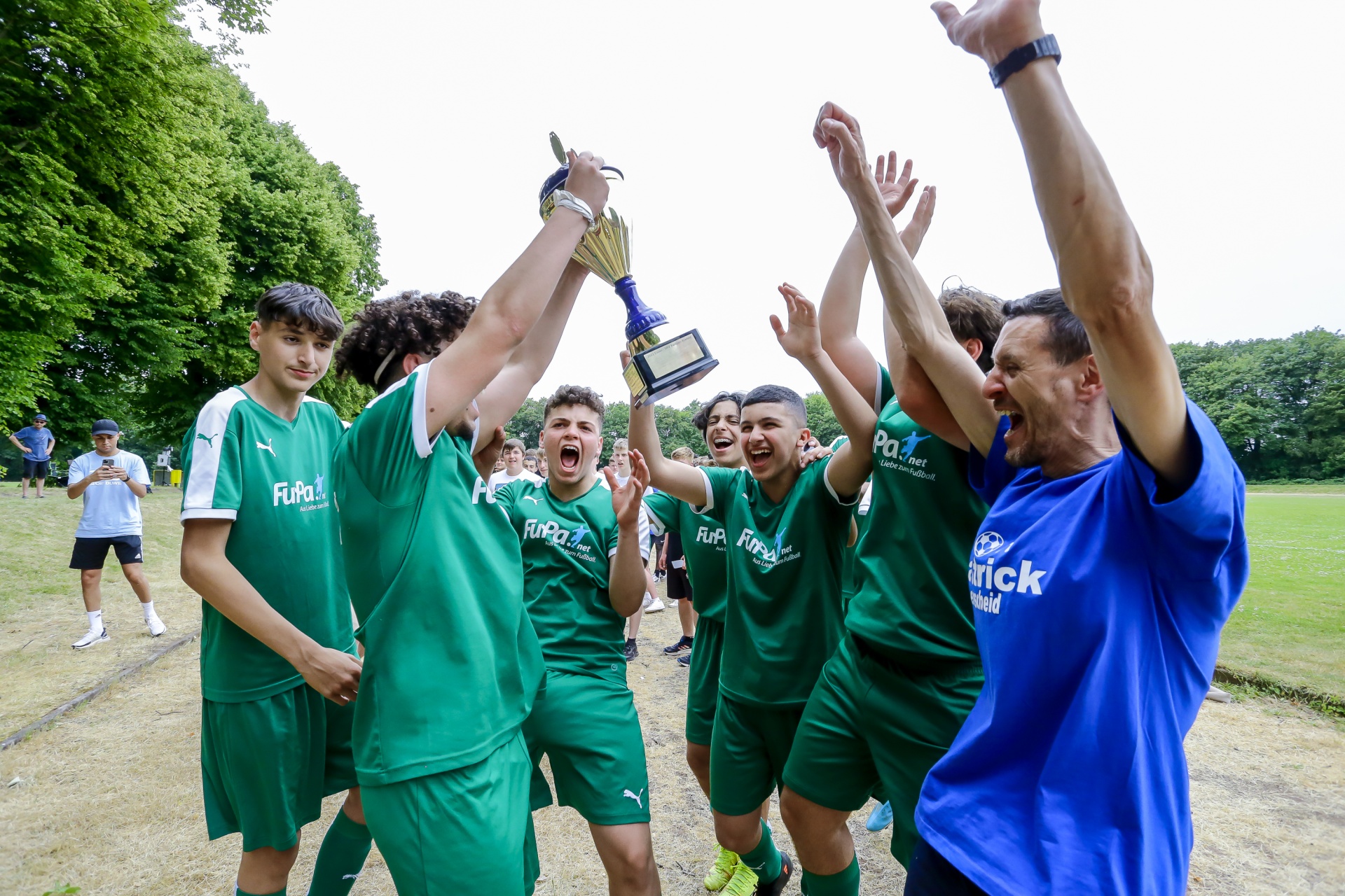 [{"clang_id":1,"value":"Fußballteam in grünen Trikots feiert Sieg mit Pokal. Jubelnde Spieler umringt von Betreuer, Hintergrund: Grünfläche und Zuschauer."}]