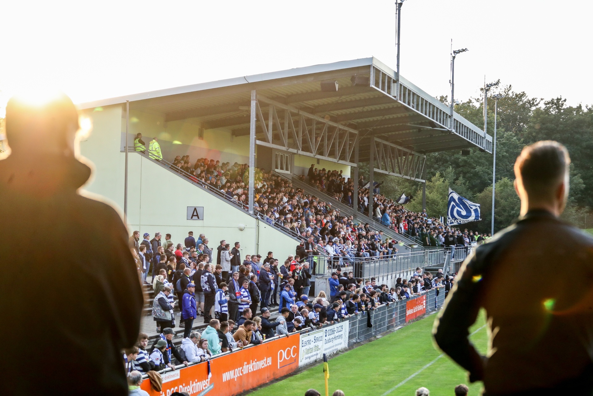 [{"clang_id":1,"value":"Fußballstadion mit Zuschauern auf den Rängen. Im Vordergrund verschwommen zwei Personen, dahinter Spielplatz und Werbebanner."}]