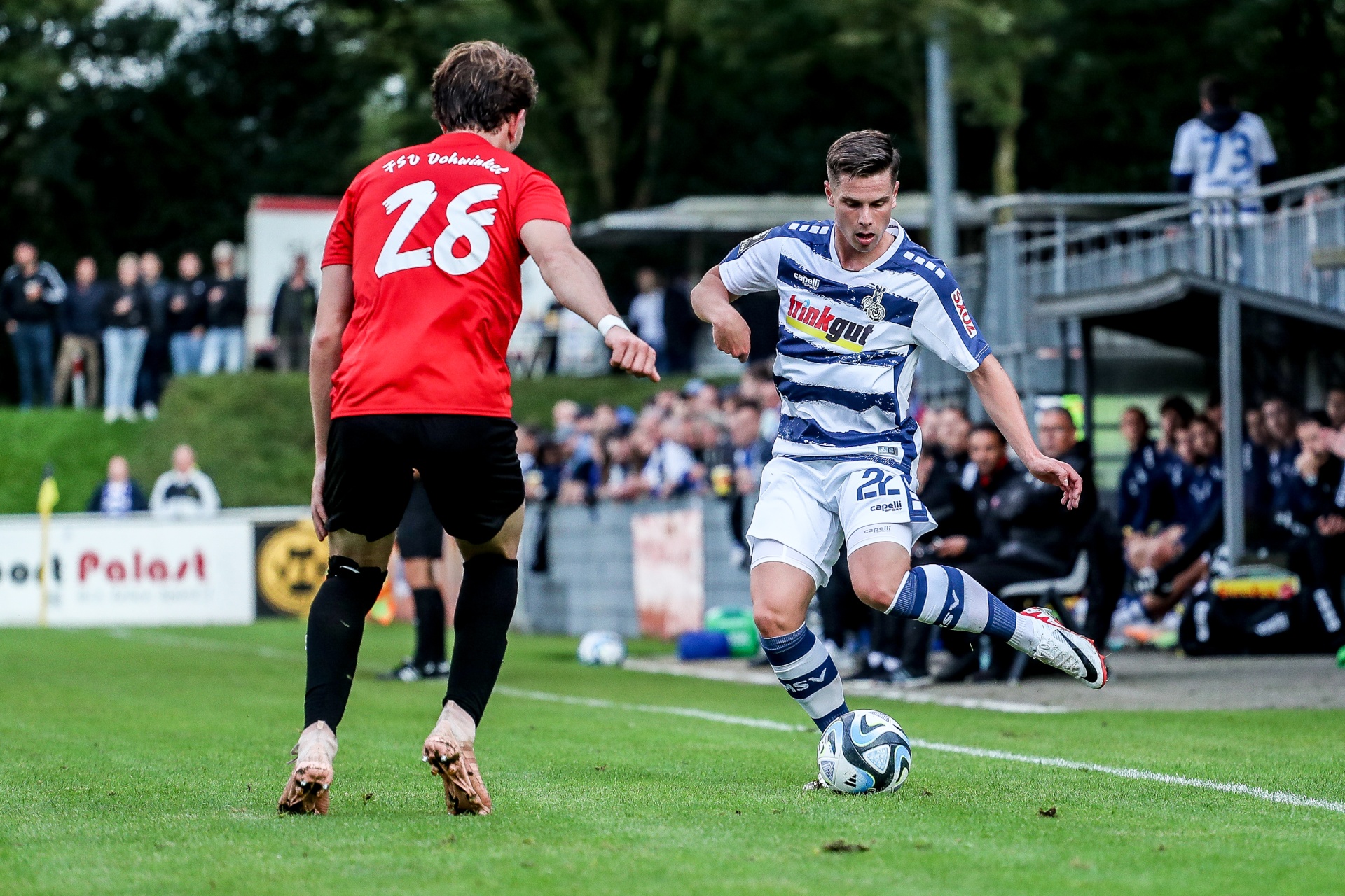 [{"clang_id":1,"value":"Zwei Fußballspieler im Spiel. Ein Spieler mit rotem Trikot und Nummer 28, der andere mit weiß-blau gestreiftem Trikot. Rasenplatz und Zuschauer im Hintergrund."}]