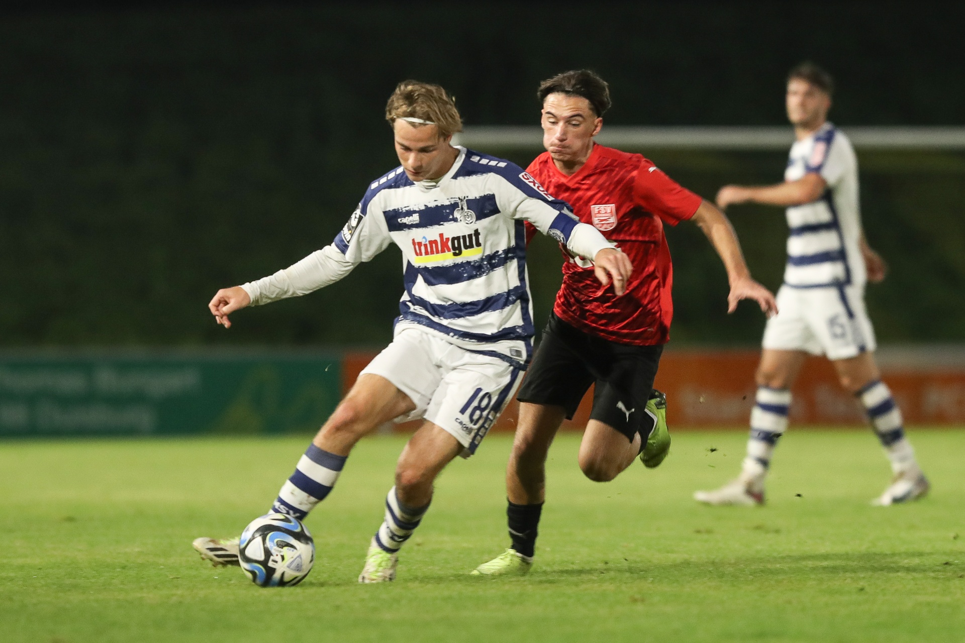 [{"clang_id":1,"value":"Zwei Fußballspieler im Spiel: Ein Spieler mit blonden Haaren und blau-weißen Streifen dribbelt den Ball, wird von einem Spieler in rotem Trikot verfolgt. Im Hintergrund ein weiterer Spieler."}]