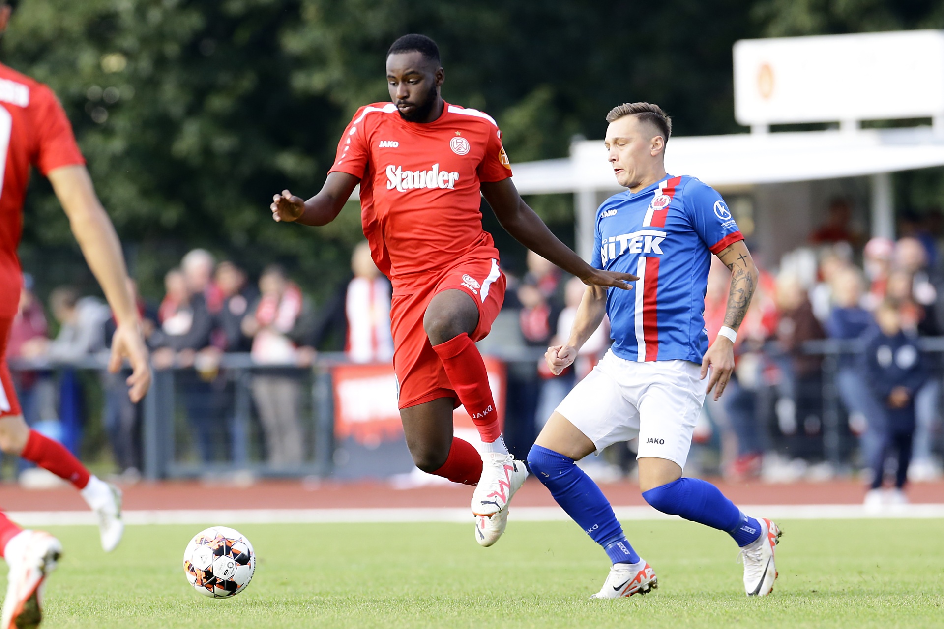 [{"clang_id":1,"value":"Zwei Fußballspieler im Wettkampf. Links ein Spieler in rotem Trikot, rechts in blau-weißem Trikot. Ein Fußball liegt auf dem Rasen vor ihnen. Im Hintergrund Zuschauer."}]