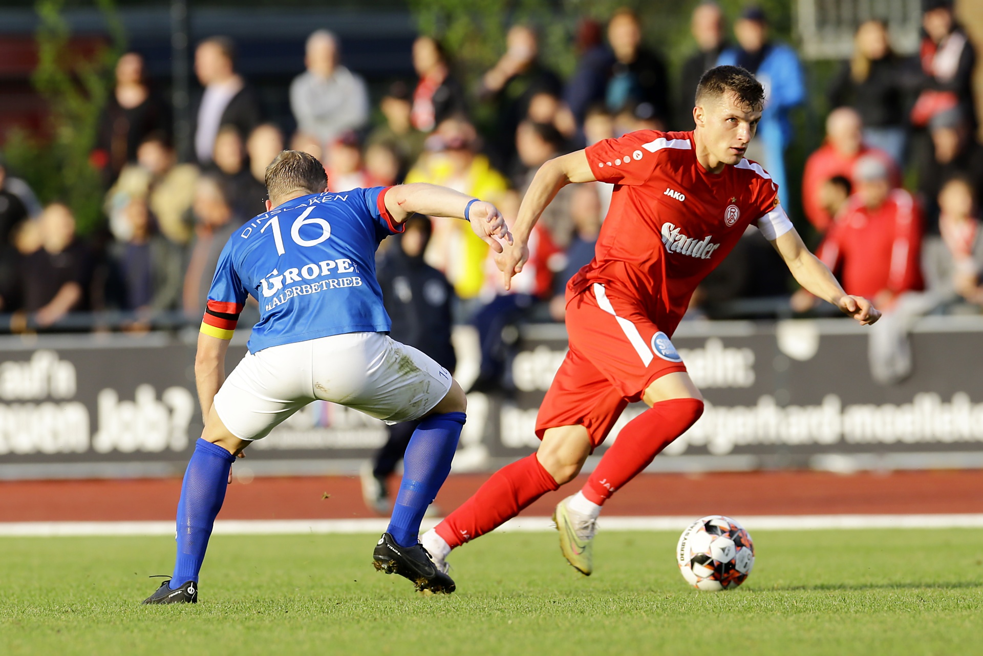 [{"clang_id":1,"value":"Zwei Fußballspieler auf grünem Rasen: Ein Spieler in Rot dribbelt den Ball, der andere in Blau-Weiß versucht, ihn zu blocken. Im Hintergrund Zuschauer."}]