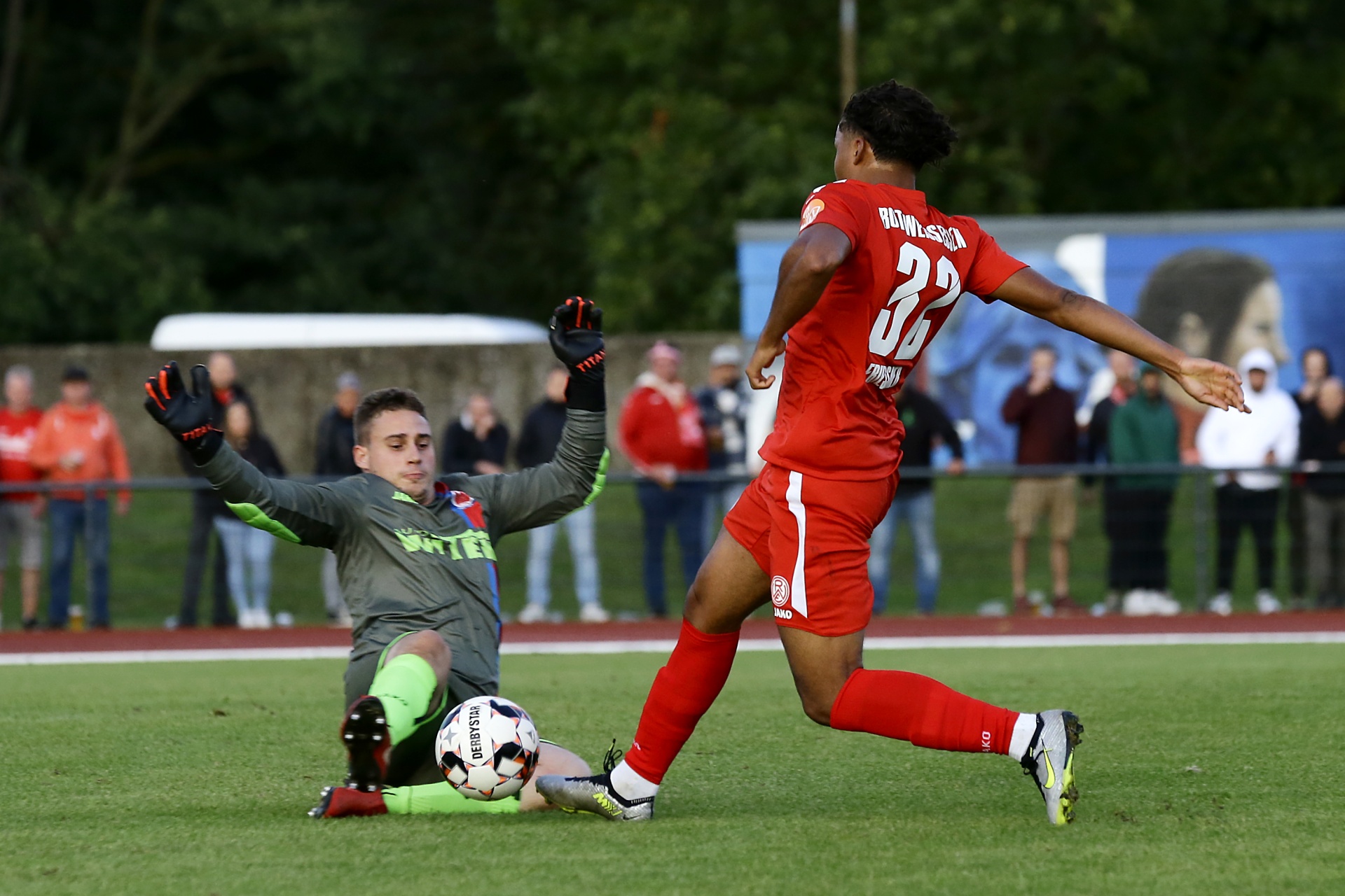 [{"clang_id":1,"value":"Fußballspiel: Spieler in Rot schießt auf das Tor, Torwart in Weiß versucht den Ball abzuwehren. Im Hintergrund Zuschauer und Bäume."}]