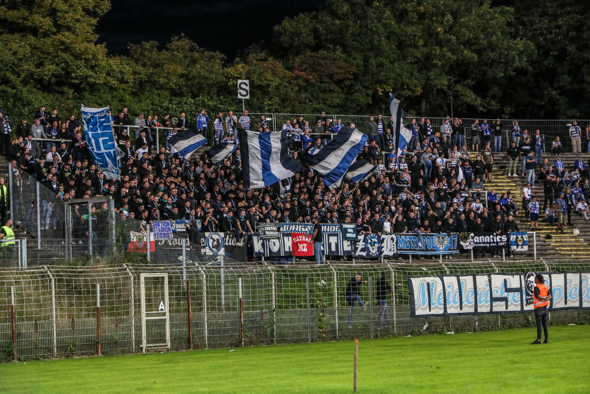 [{"clang_id":1,"value":"Fußballstadion: Fans mit blauen und weißen Fahnen auf der Tribüne. Im Hintergrund Hügel und dunkler Himmel. Spielfeld im Vordergrund."}]