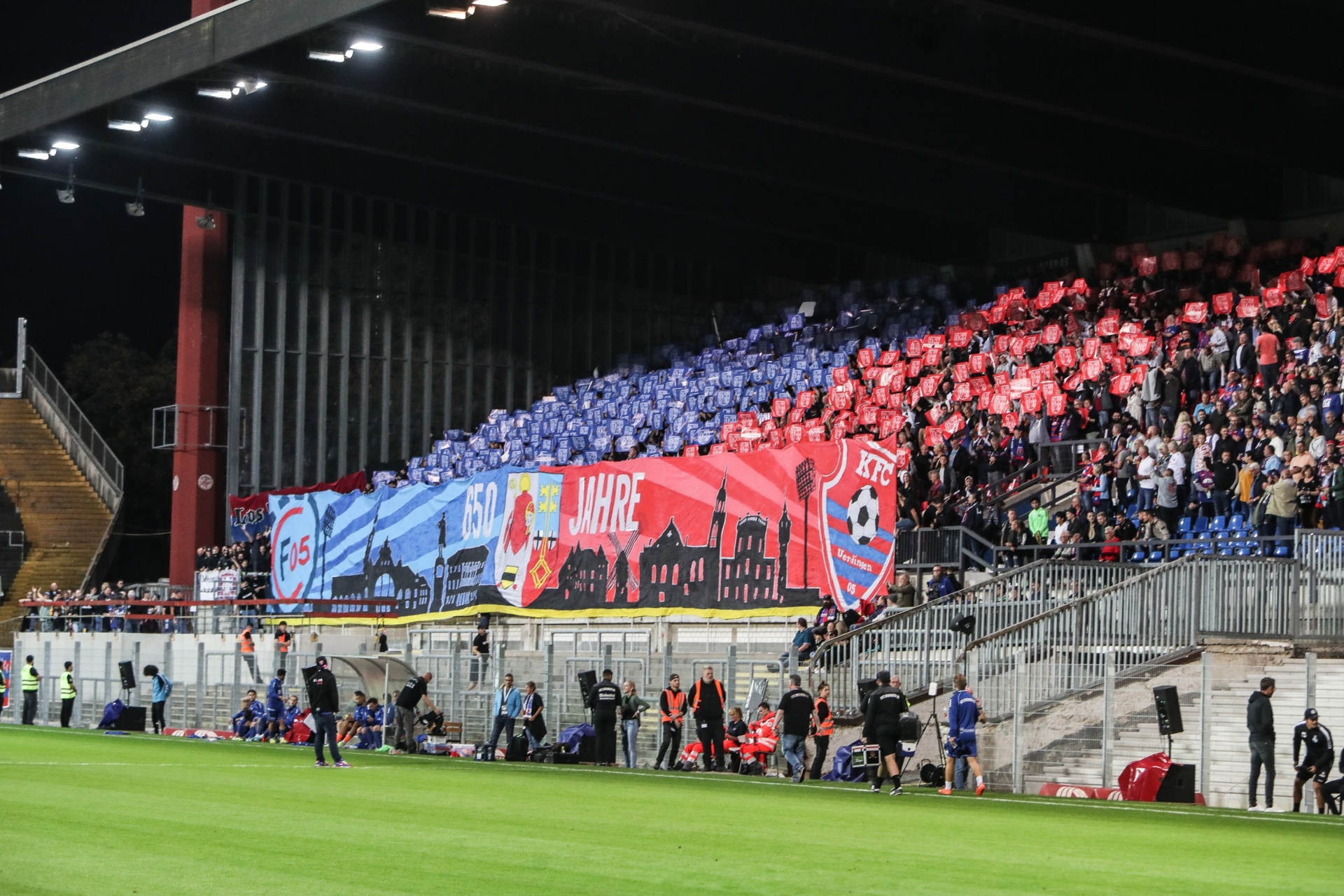 [{"clang_id":1,"value":"Fußballstadion bei Nacht: Volle Zuschauertribünen in Rot und Blau. Großes Banner mit Schrift und Skyline im Hintergrund. Rasen im Vordergrund."}]