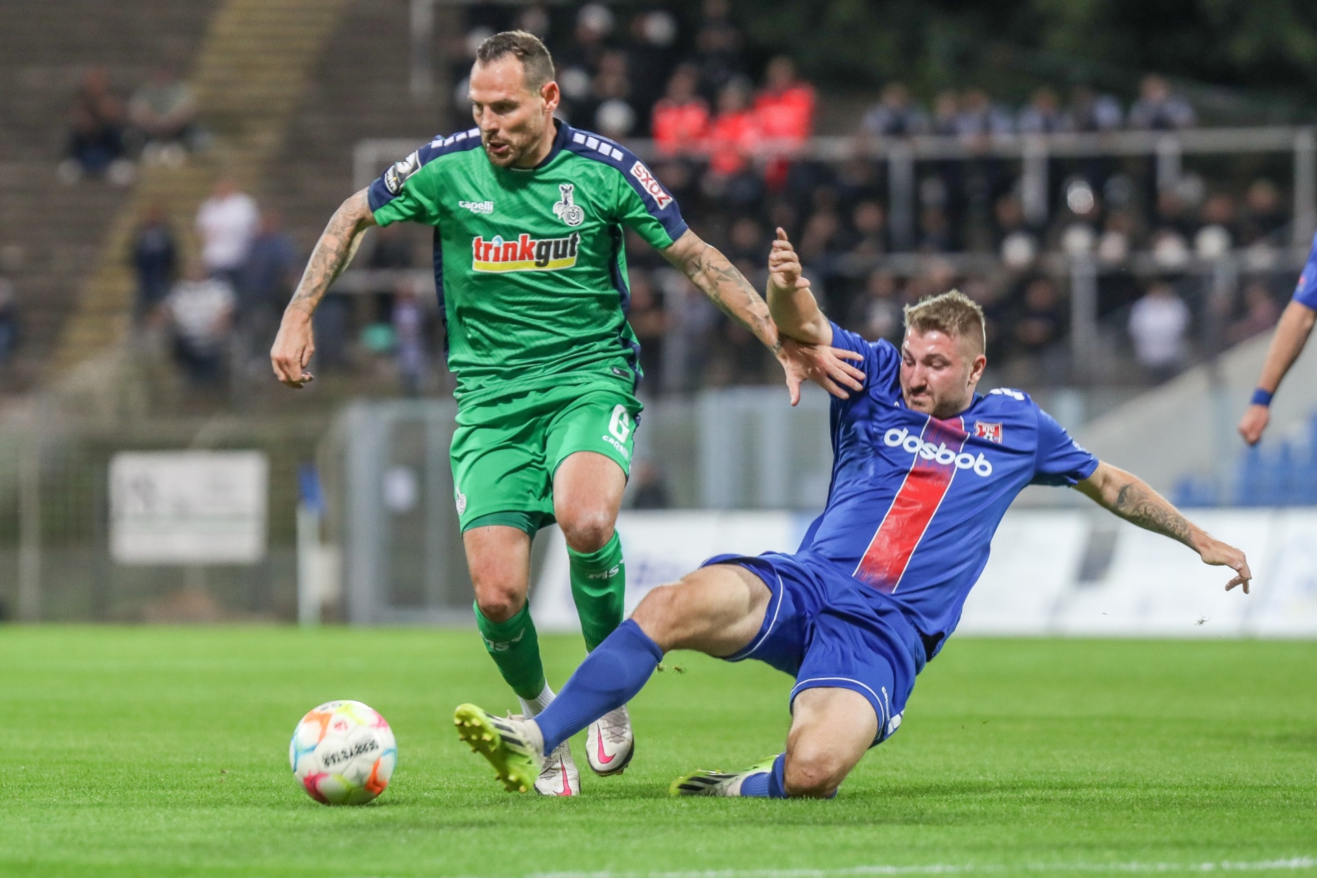 [{"clang_id":1,"value":"Zwei Fußballspieler im Spiel. Links ein Spieler in grünem Trikot, rechts ein Spieler in blau-rotem Trikot rutscht. Im Hintergrund Zuschauer und Stadion."}]