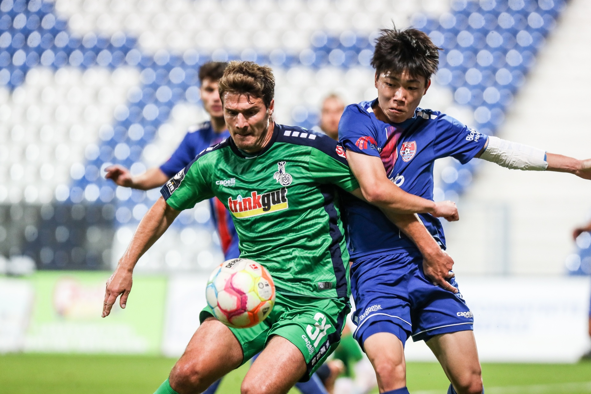 [{"clang_id":1,"value":"Fußballspieler im Zweikampf. Links ein Spieler in grünem Trikot mit Ball, rechts ein Spieler in blauem Trikot. Im Hintergrund Zuschauer auf den Tribünen."}]