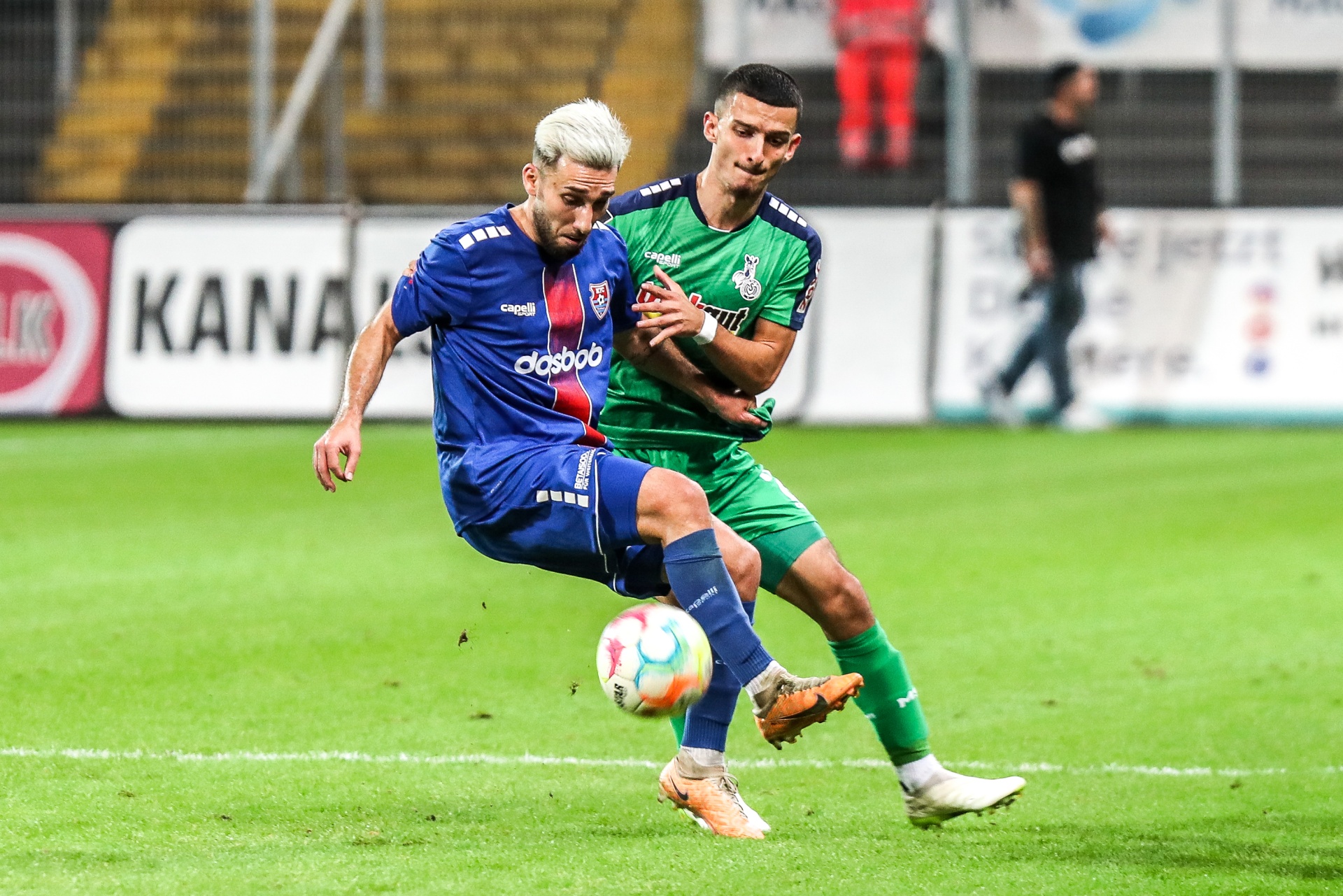 [{"clang_id":1,"value":"Zwei Fußballspieler im Wettkampf auf grünem Rasen. Links ein Spieler mit blauem Trikot und weiß-blond gefärbten Haaren beim Schießen, rechts ein Spieler mit grünem Trikot. Im Hintergrund Zuschauer und Werbebanner."}]