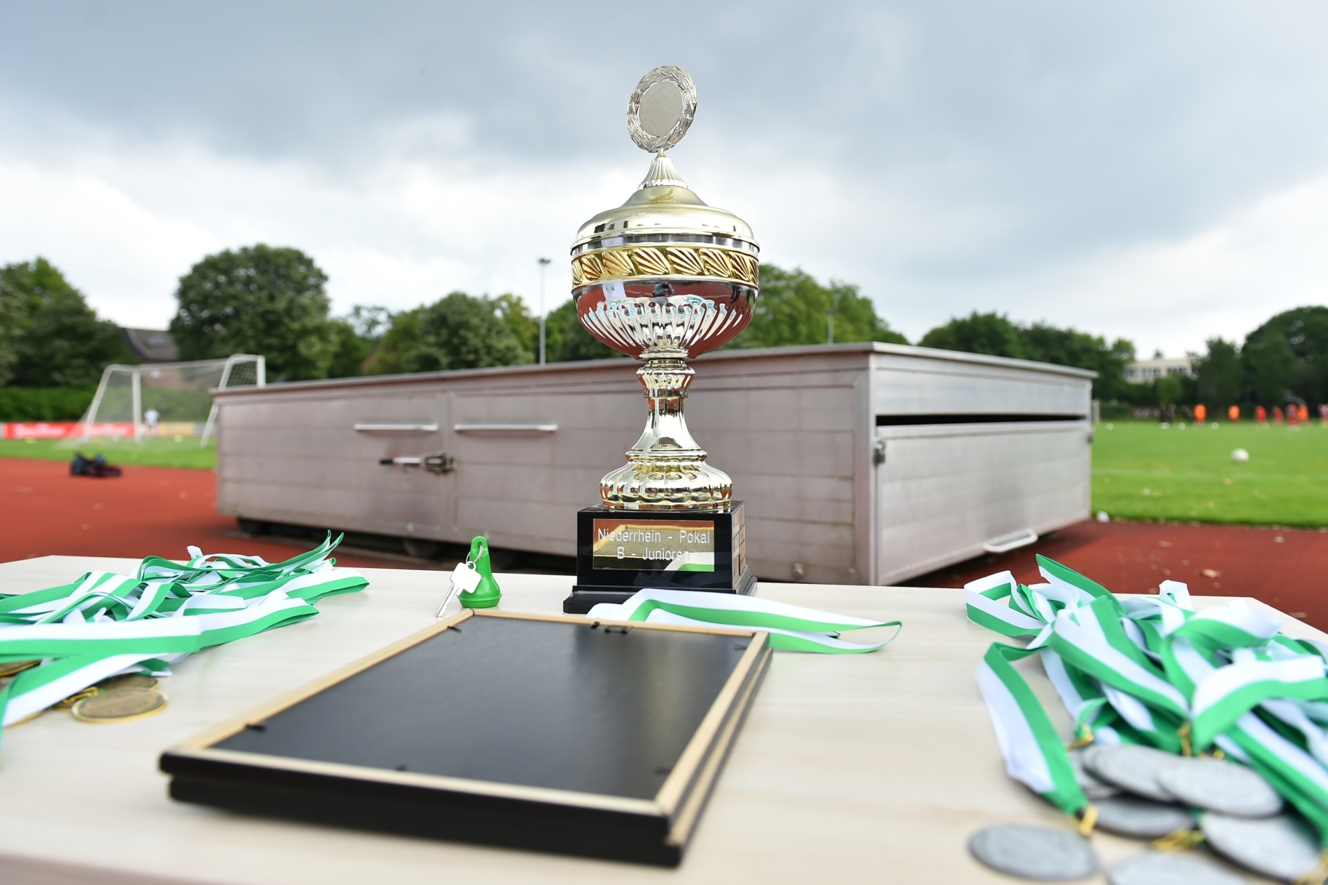 [{"clang_id":1,"value":"Pokal und Urkunde auf Tisch im Freien. Im Hintergrund ein Fußballfeld und Gebäude unter bewölktem Himmel. Grüne und weiße Bänder liegen verstreut."}]