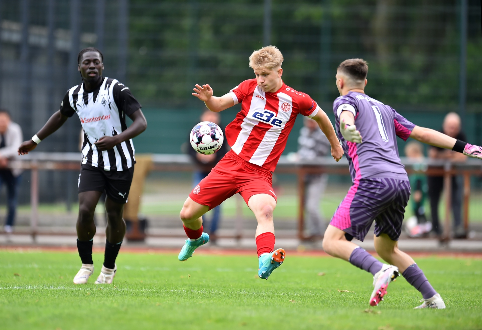 [{"clang_id":1,"value":"Fußballspiel. Spieler mit rotem Trikot dribbelt Ball, Gegner in lila und schwarz-weiß im Hintergrund. Grüne Rasenfläche, Zaun im Hintergrund."}]
