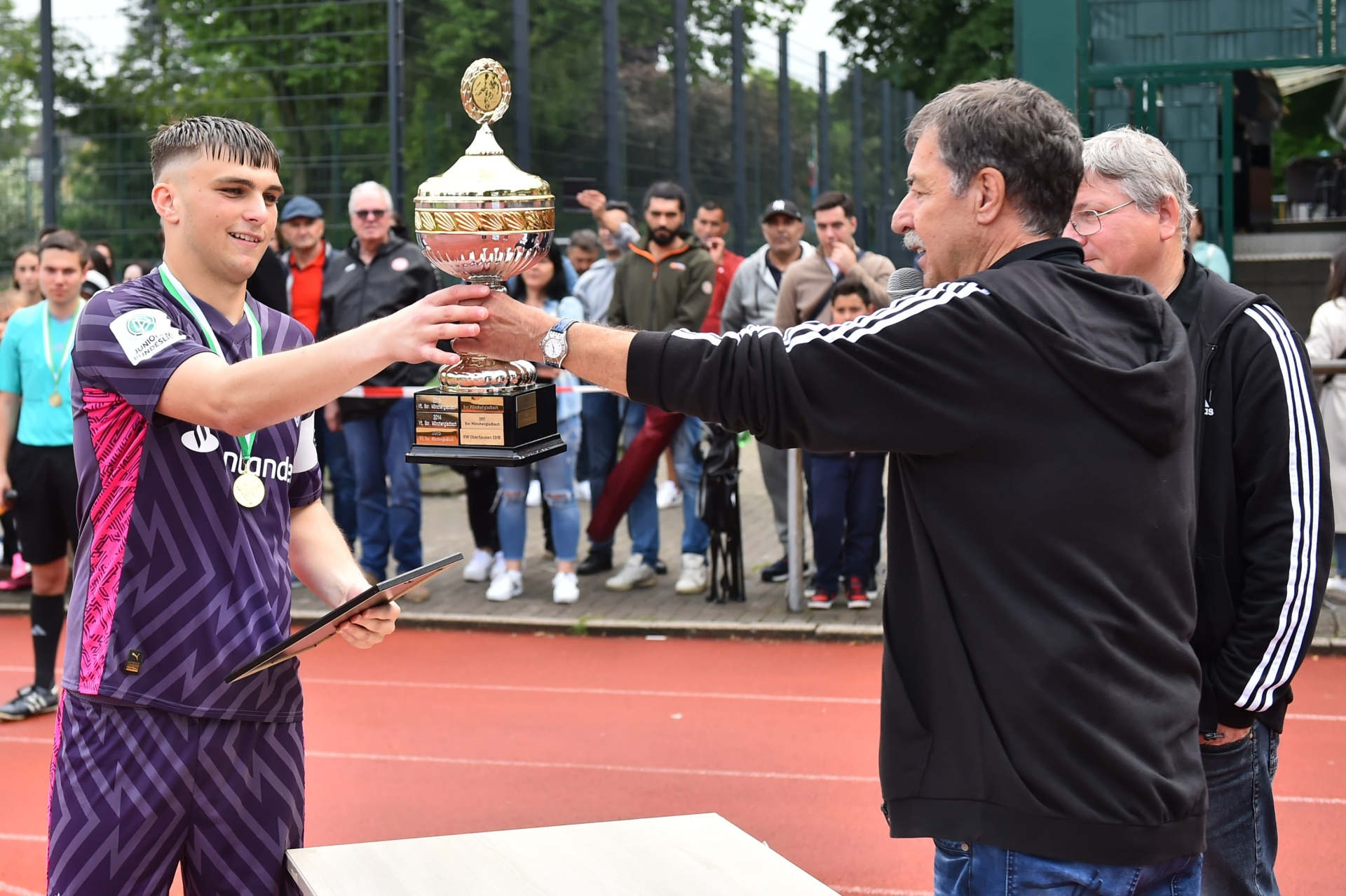 [{"clang_id":1,"value":"Ein Fußballspieler in Vereinskleidung erhält von einem Mann einen Pokal auf einem roten Untergrund. Im Hintergrund stehen weitere Personen und ein Tennisplatz."}]