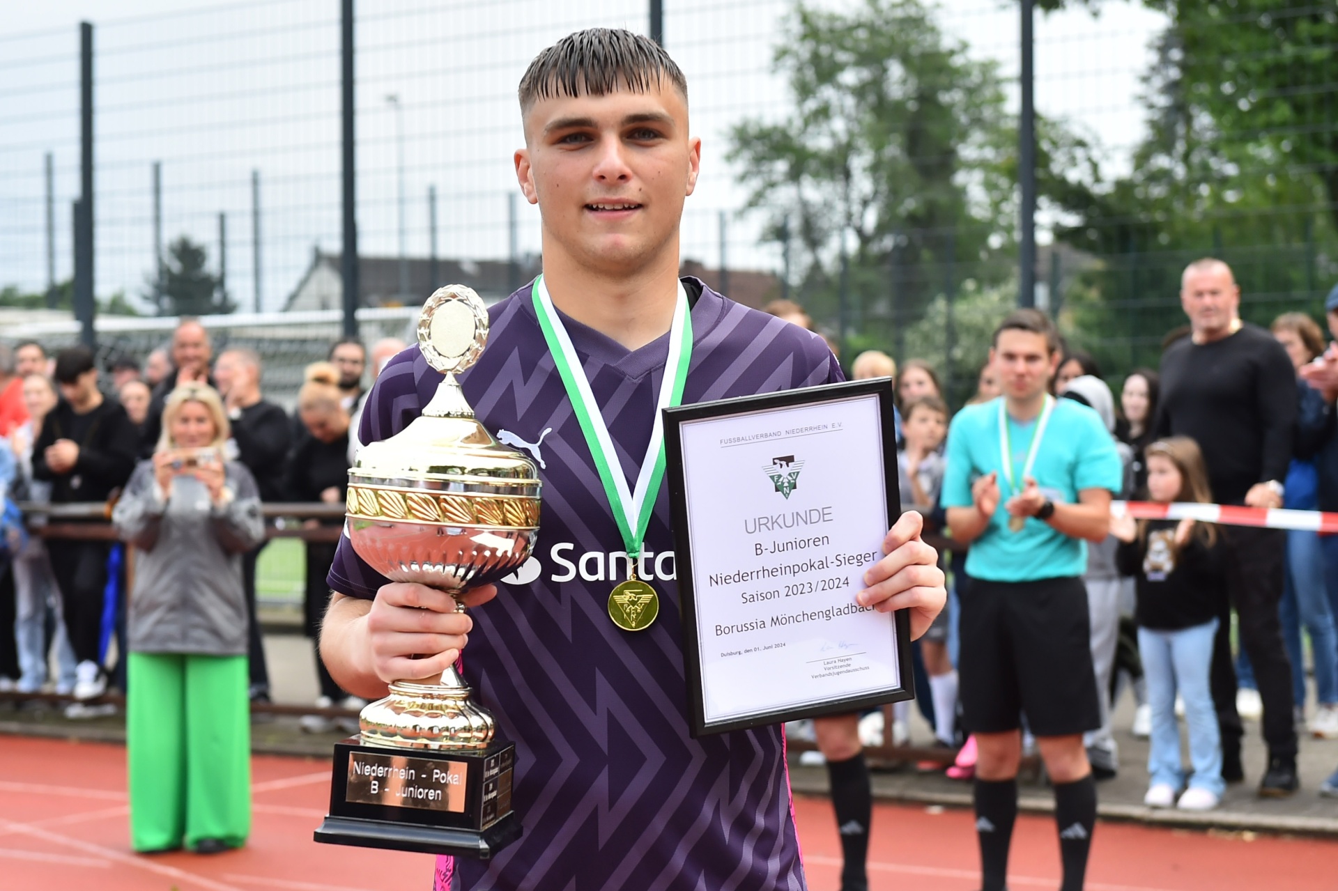 [{"clang_id":1,"value":"Junger Fussballspieler mit Trikot hält Pokal und Urkunde. Im Hintergrund weitere Personen und grüne Bäume."}]