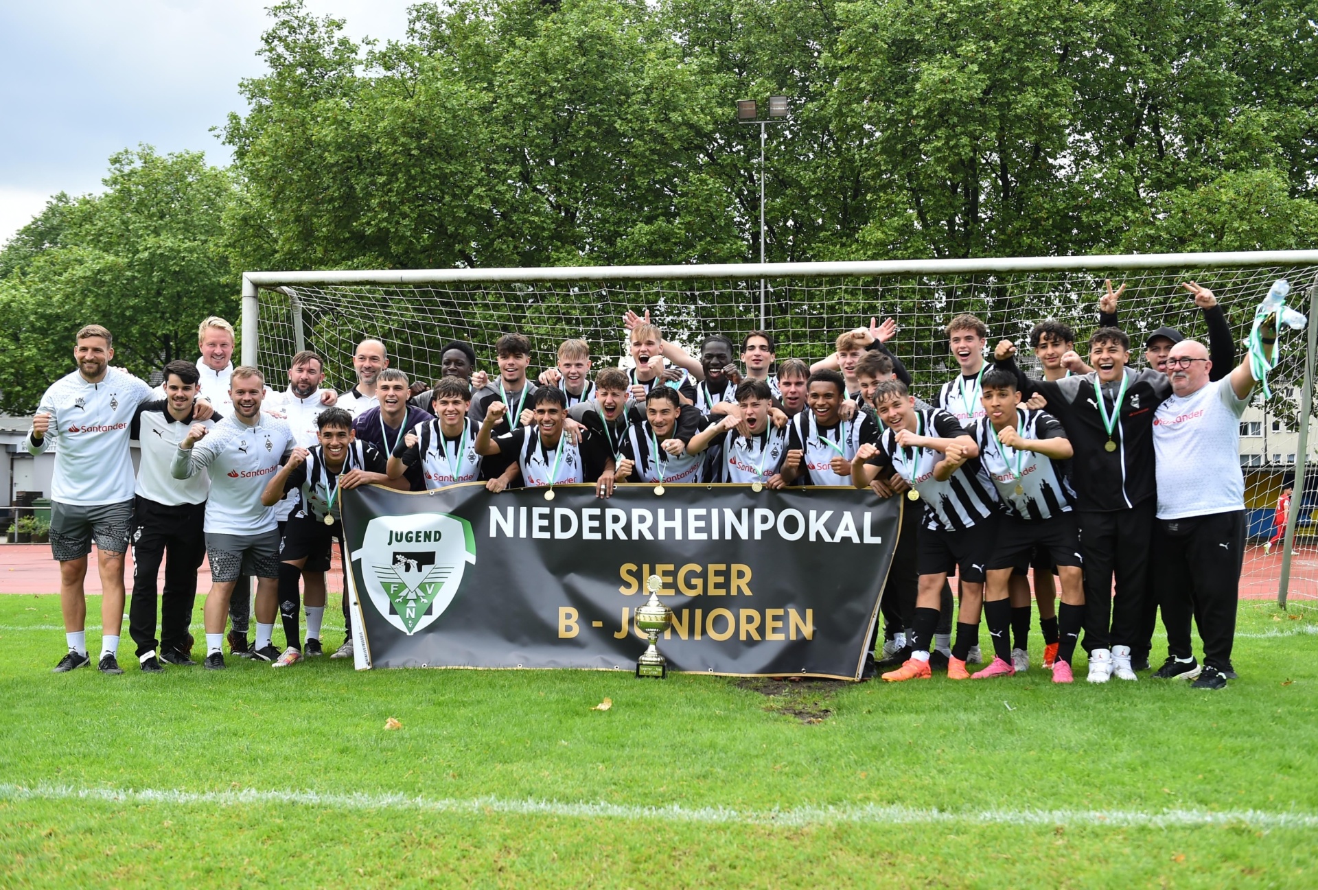 [{"clang_id":1,"value":"Fußballmannschaft mit Banner \"Niederrheinpokal Sieger B-Junioren\" auf grünem Rasen vor Fußballtor. Trainer und Spieler feiern, einige heben Pokal und grüne Fahne hoch."}]