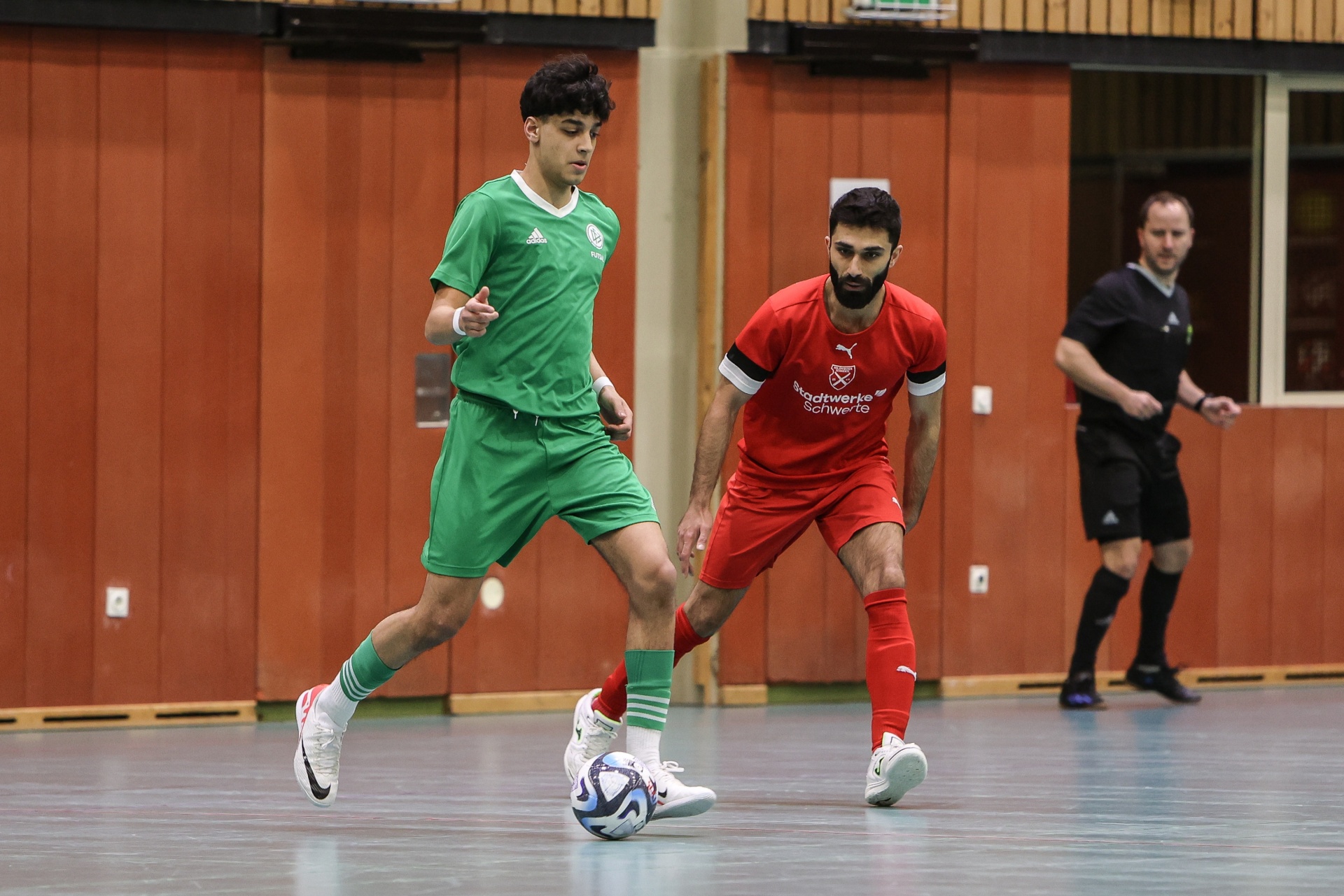 [{"clang_id":1,"value":"Zwei Fußballspieler in Hallenfußball-Montage: links grün, rechts rot. Im Hintergrund eine Person in Schwarz. Holzwand im Hintergrund."}]
