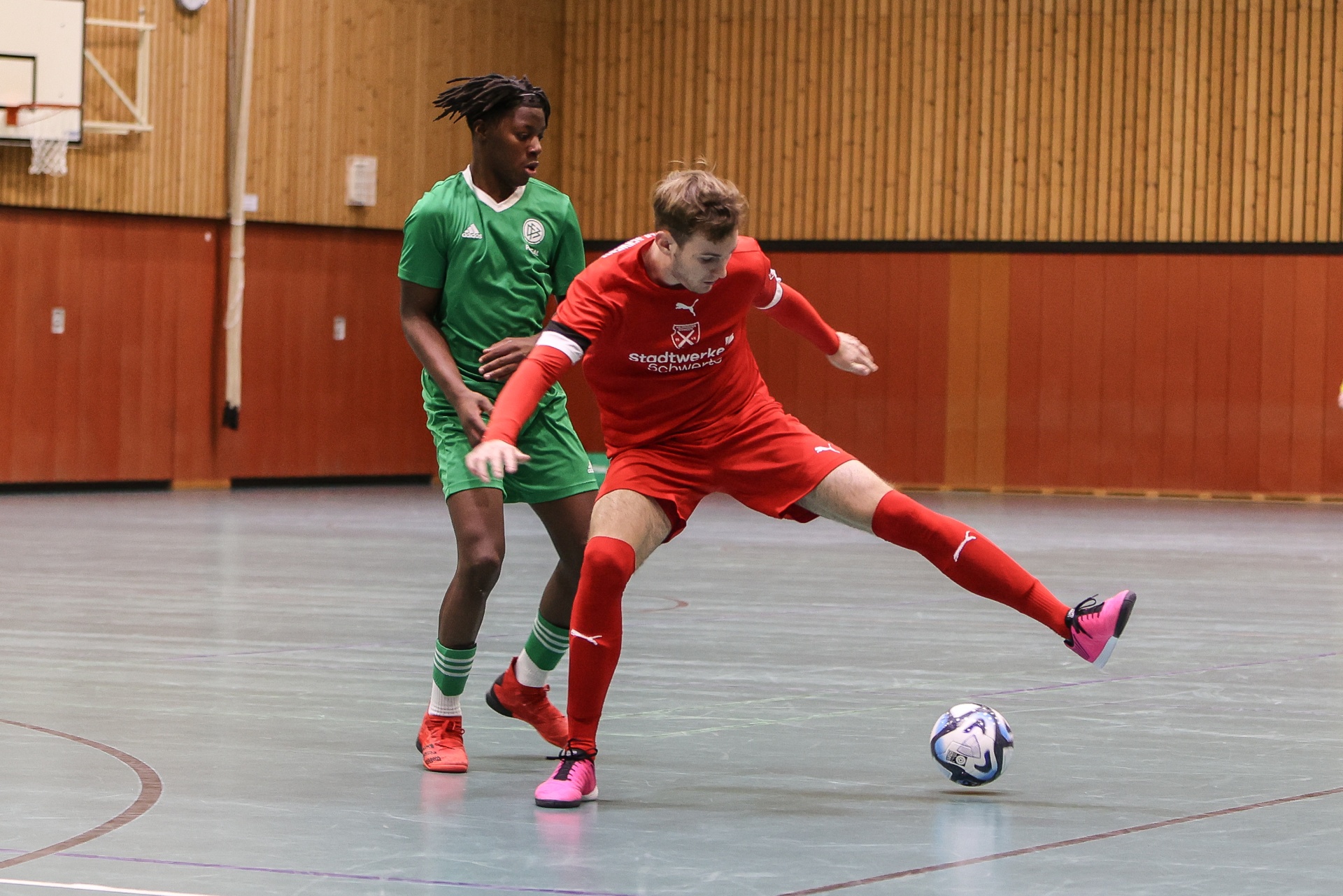 [{"clang_id":1,"value":"Zwei Fußballspieler in Sporthalle. Links dunkelhäutiger Spieler in grünem Trikot, rechts hellhäutiger Spieler in rotem Trikot, der Ball spielt."}]