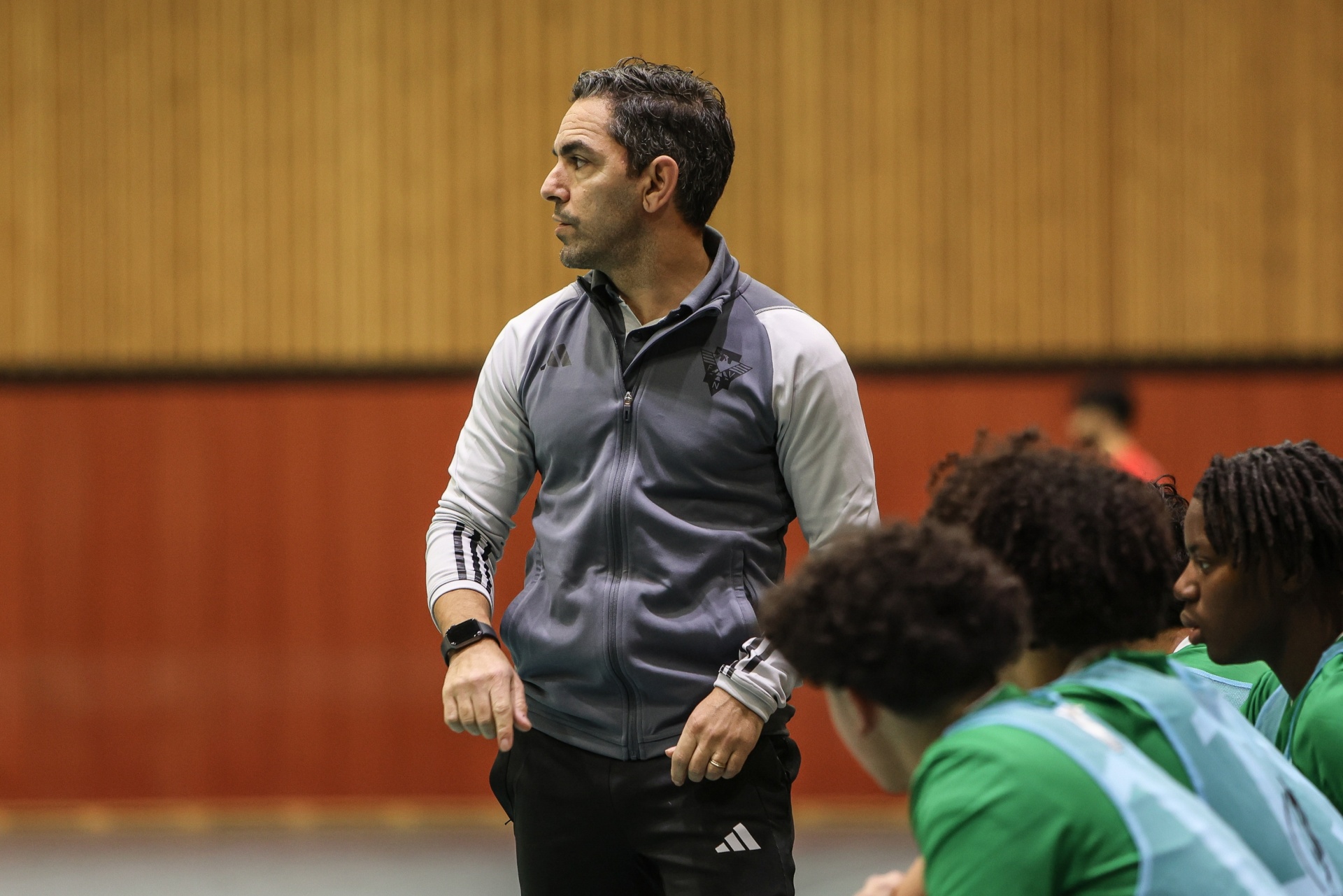 [{"clang_id":1,"value":"Mann mit Adidas-Trainingsjacke steht in einer Sporthalle, blickt auf eine Gruppe Basketballspieler mit lockigem Haar und grünen Trikots."}]