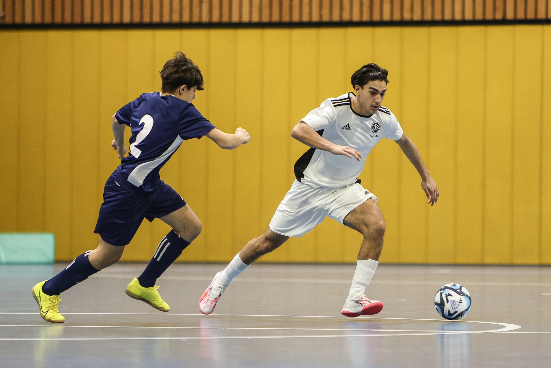 [{"clang_id":1,"value":"Zwei Futsalspieler in Hallensportarena. Ein Spieler in weiß dribbelt den Ball, während ein Gegner in blau ihn verfolgt. Gelbe Wand im Hintergrund."}]