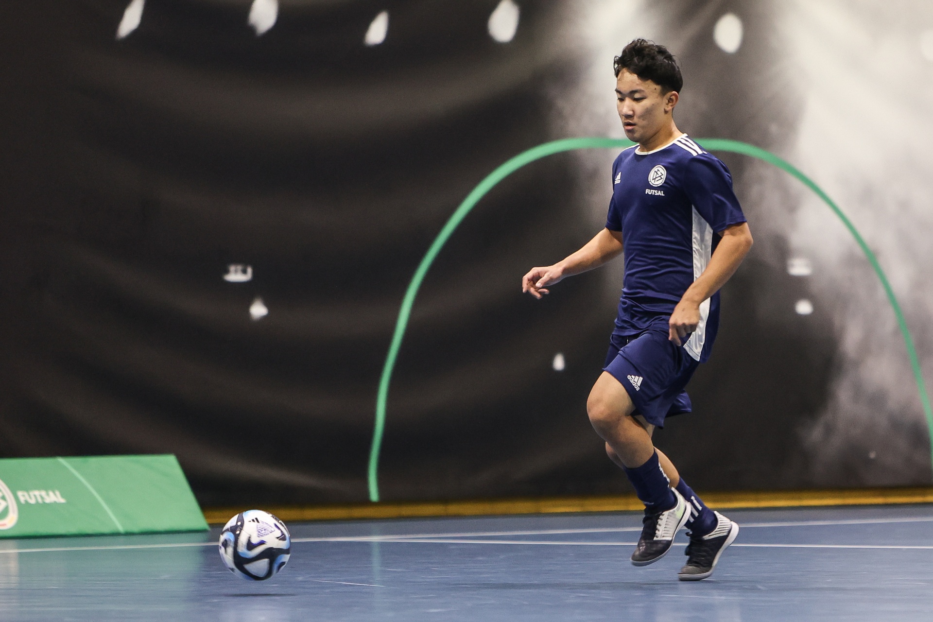 [{"clang_id":1,"value":"Futsalspieler dribbelt den Ball auf blauem Hallenboden. Er trägt ein dunkelblaues Trikot und Shorts mit weißen Adidas-Streifen. Im Hintergrund eine grüne Wand mit Futsal-Logo."}]