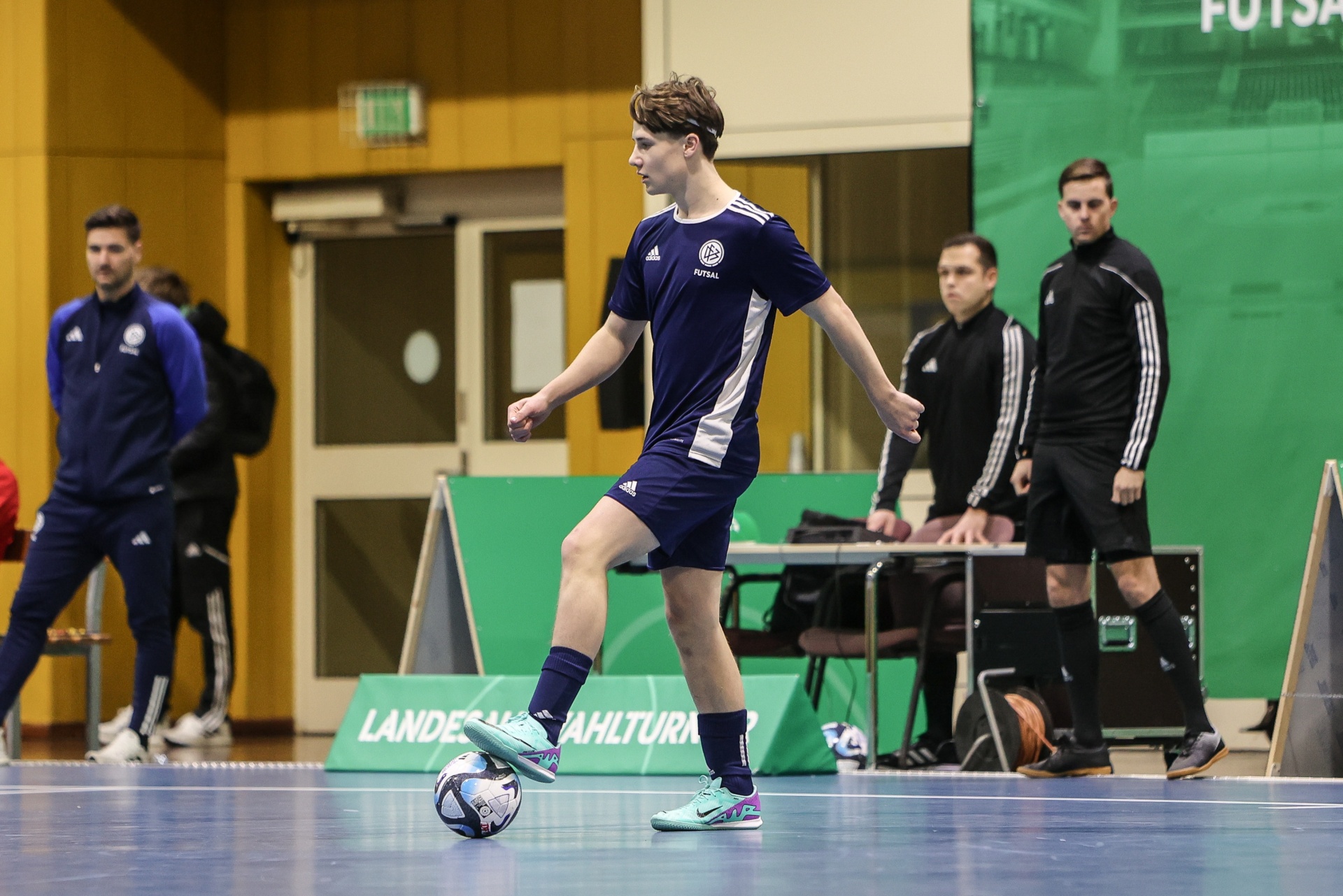 [{"clang_id":1,"value":"Futsalspieler mit blauem Trikot dribbelt Ball in Sporthalle. Im Hintergrund Zuschauer und Begrenzung durch Werbebanner."}]