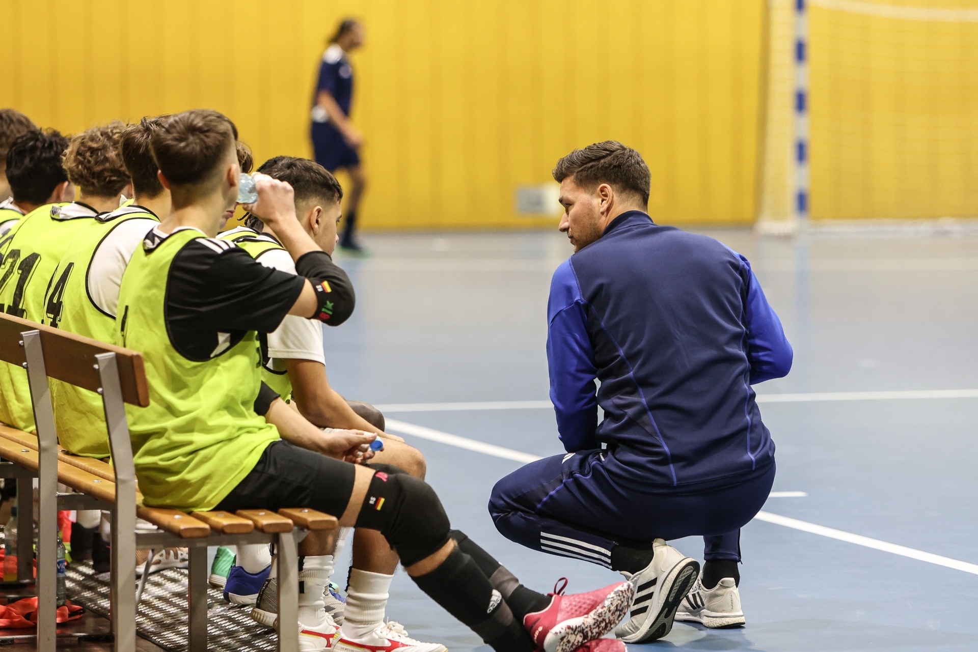 [{"clang_id":1,"value":"Handballtrainer in blauem Trainingsanzug in der Halle. Er bespricht Taktik mit Spielern auf der Bank. Einige Spieler tragen grüne Westen."}]