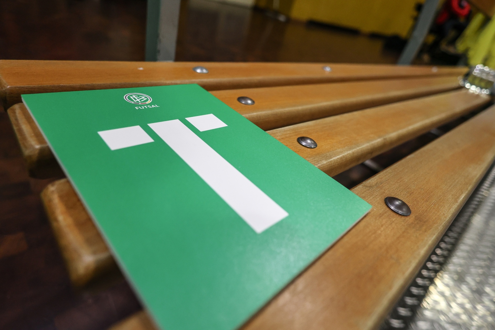 [{"clang_id":1,"value":"Grüner Futsal-Spielplan liegt auf einer hölzernen Bank mit Metallbeschlägen. Im Hintergrund verschwommene Sporthalle."}]
