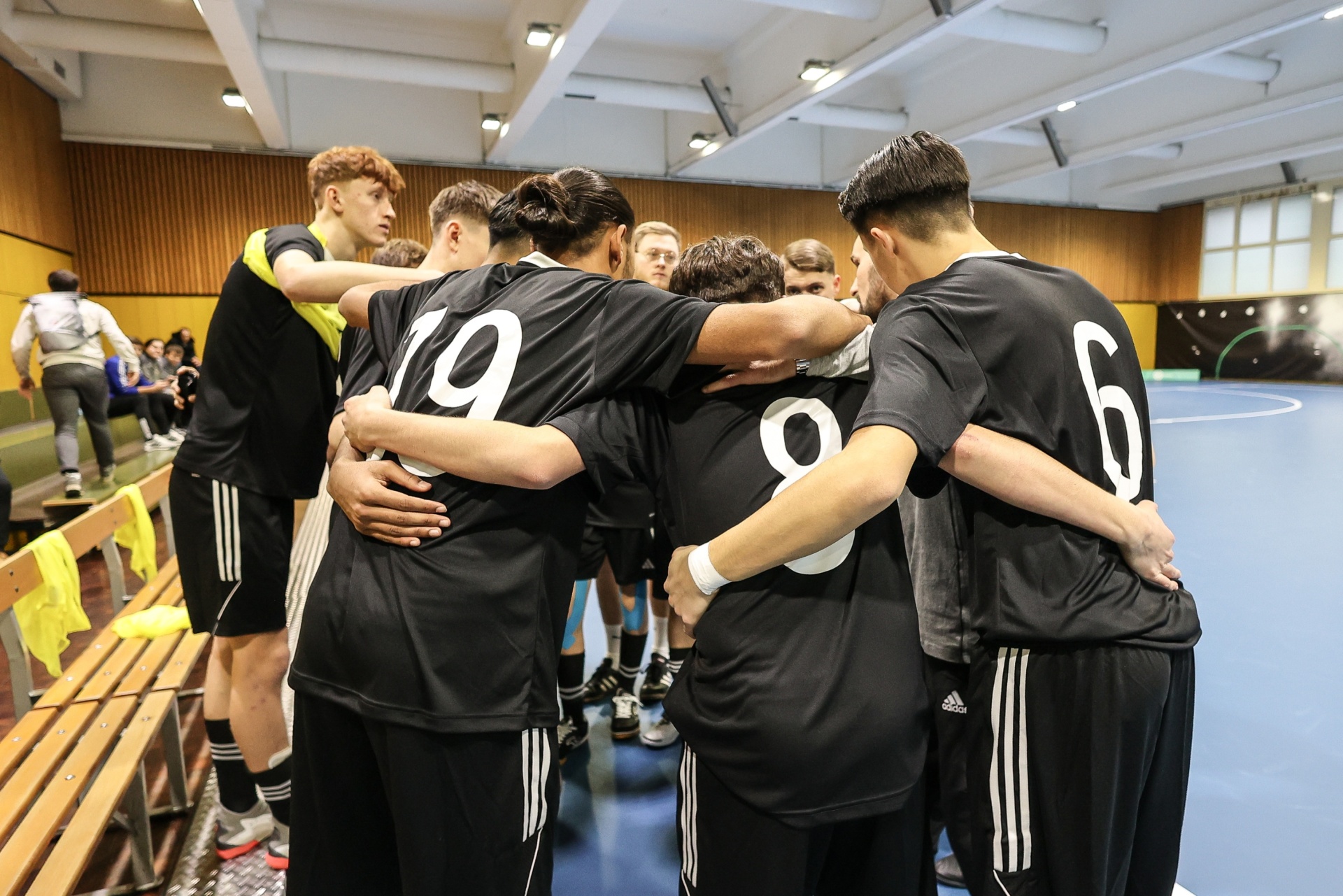 [{"clang_id":1,"value":"Futsalteam umringt sich zum Gespräch. Spieler tragen schwarze Trikots mit weißen Nummern (6, 9). Im Hintergrund Sporthalle mit Bänken und Zuschauern."}]