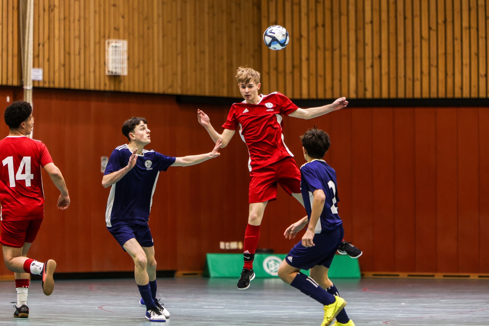 [{"clang_id":1,"value":"Fußballspiel in Sporthalle. Ein Spieler in Rot springt und köpft den Ball. Zwei weitere Spieler in Blau und Rot stehen darum. Holzvertäfelung im Hintergrund."}]