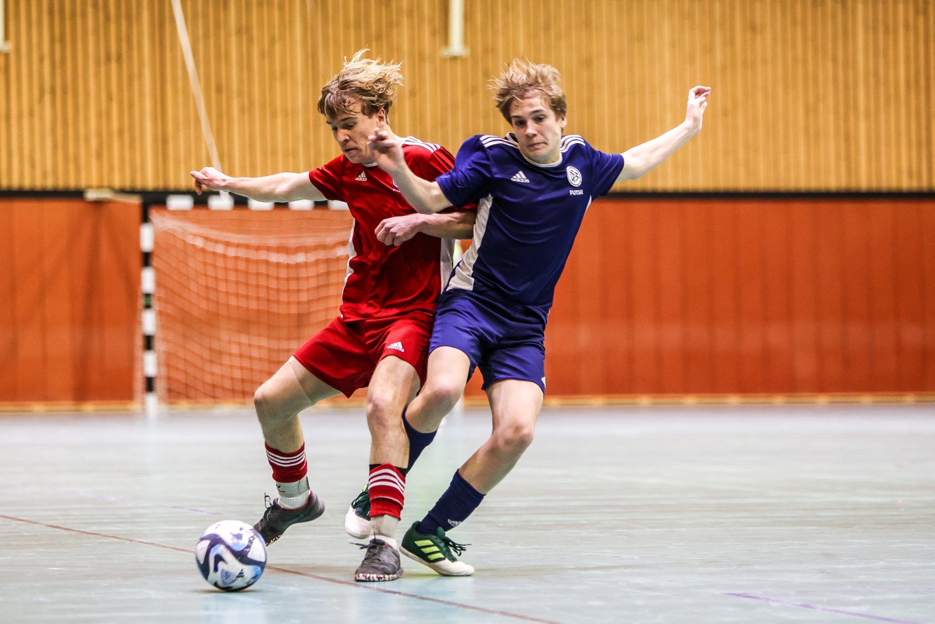[{"clang_id":1,"value":"Zwei junge Fussballspieler mit langen, blonden Haaren stehen sich auf einem Hallenfußballplatz gegenüber. Links trägt der Spieler Rot, rechts Blau. Ein Fussball liegt zwischen ihnen."}]