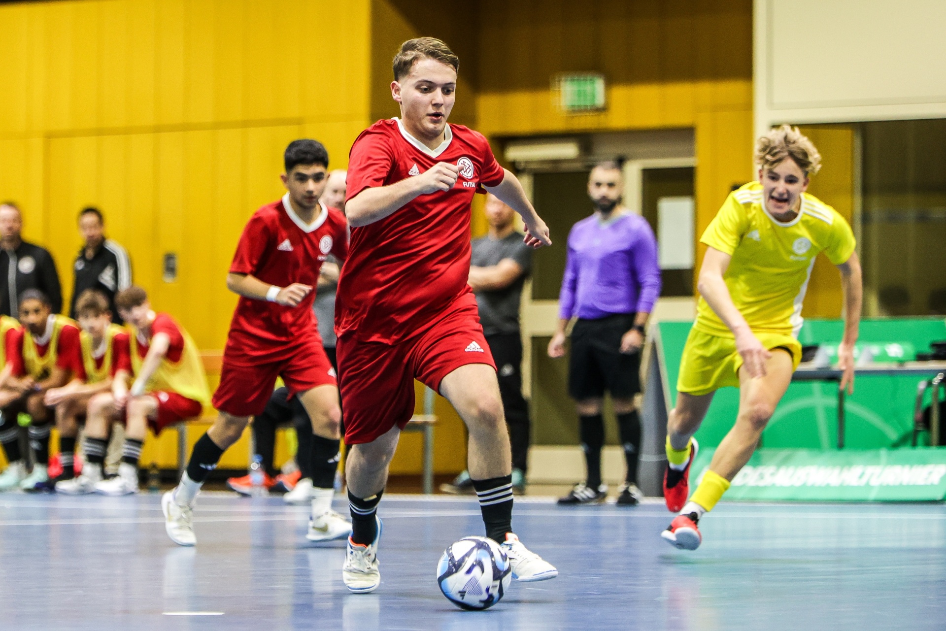 [{"clang_id":1,"value":"Futsalspiel in Sporthalle. Spieler mit rotem Trikot dribbelt Ball, Gegenspieler in gelbem Trikot im Hintergrund. Schiedsrichter erkennbar."}]