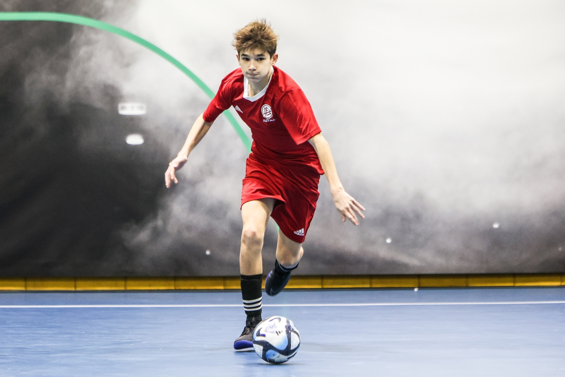 [{"clang_id":1,"value":"Junge in rotem Fußballtrikot dribbelt mit Ball auf blauem Hallenboden. Adidas-Logo auf Trikot und Schuh. Im Hintergrund graue Wand mit grüner Linie."}]