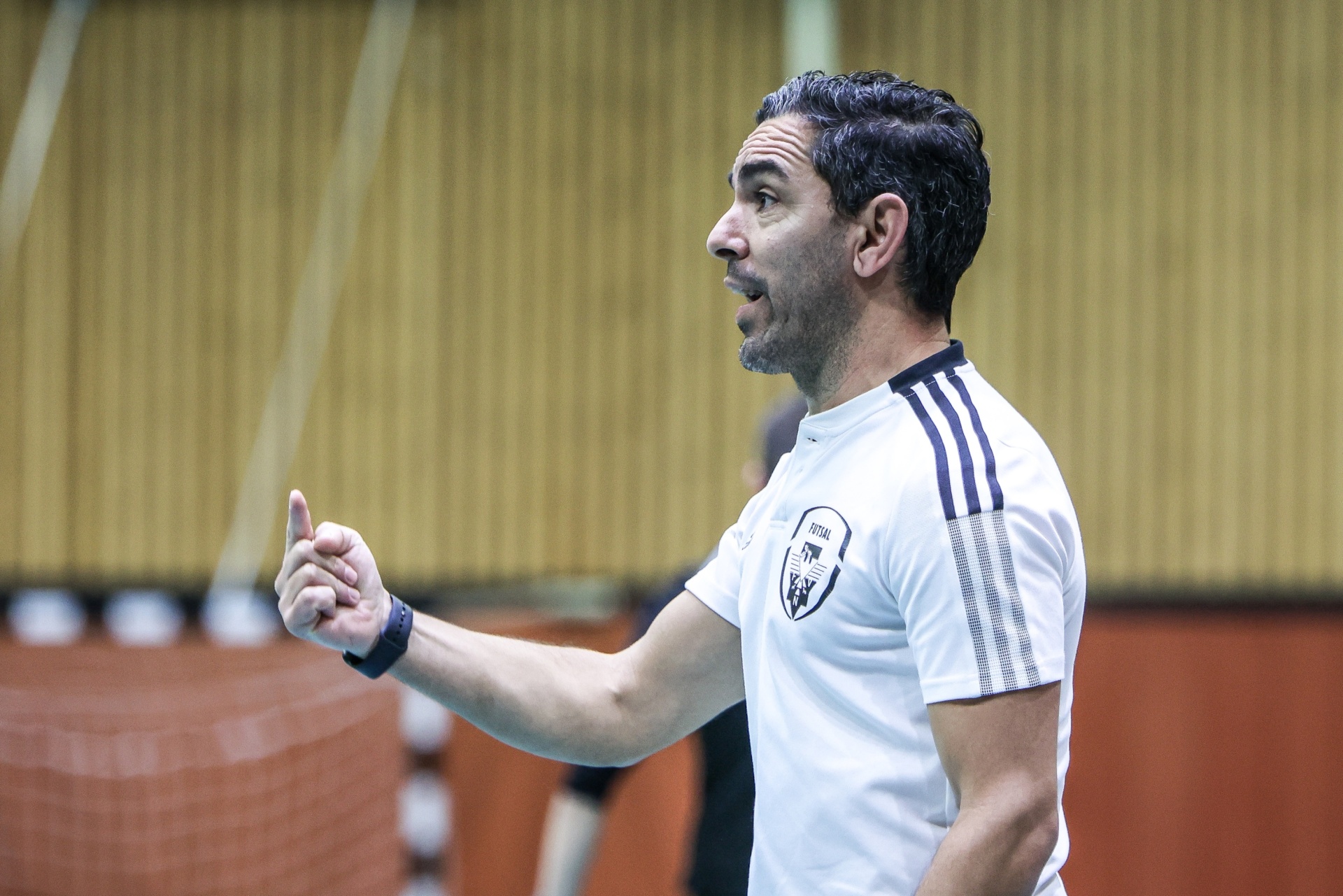 [{"clang_id":1,"value":"Trainer mit grau meliertem Haar in weißem Polo mit schwarzem Logo, gestikulierend mit erhobenem Daumen. Hallenfußballtor im Hintergrund."}]