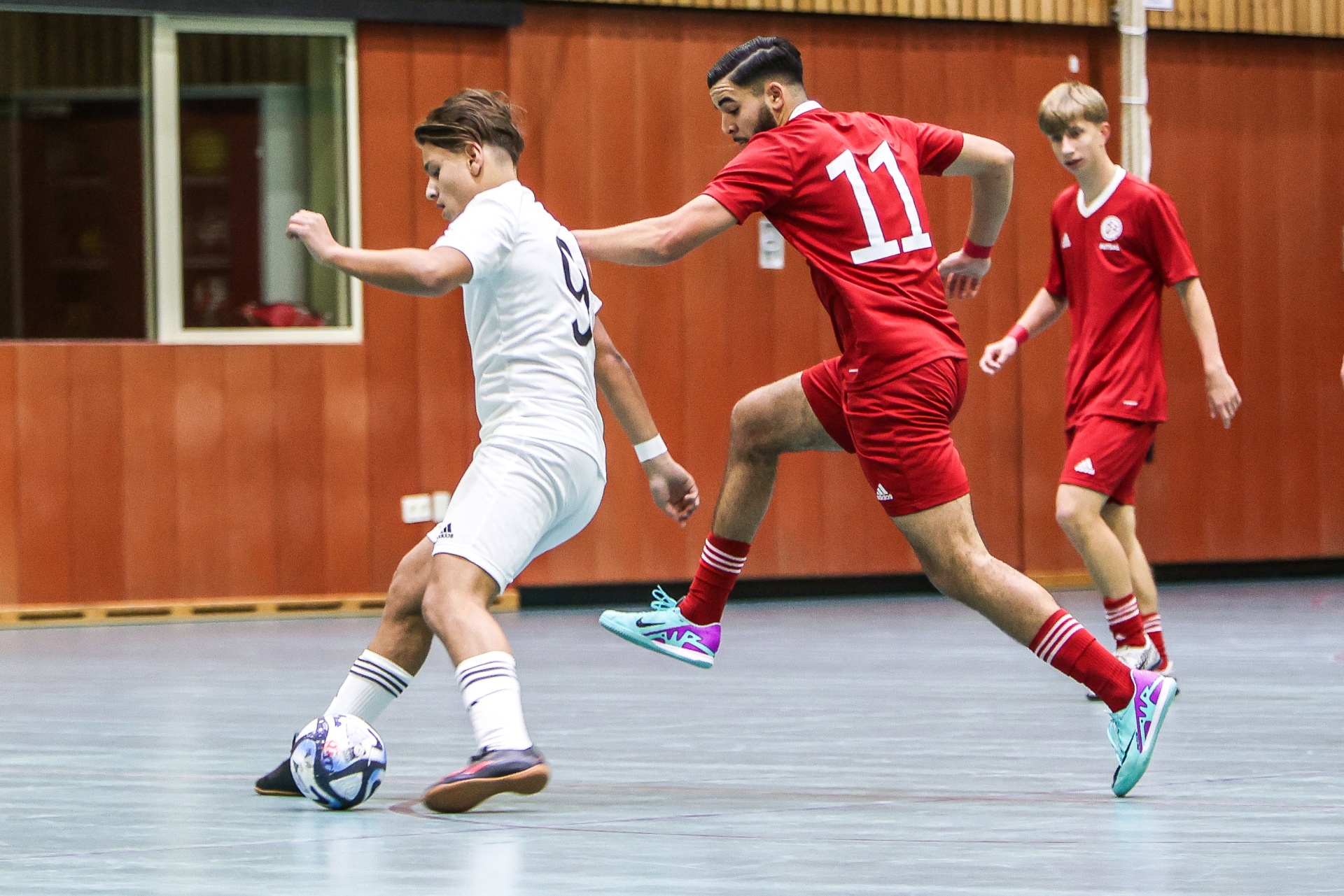 [{"clang_id":1,"value":"Futsal-Spiel: Zwei Spieler dribbeln den Ball, ein dritter im Hintergrund. Einer trägt Weiß, die anderen Rot mit Nummer 11. Holzboden, Sporthalle."}]